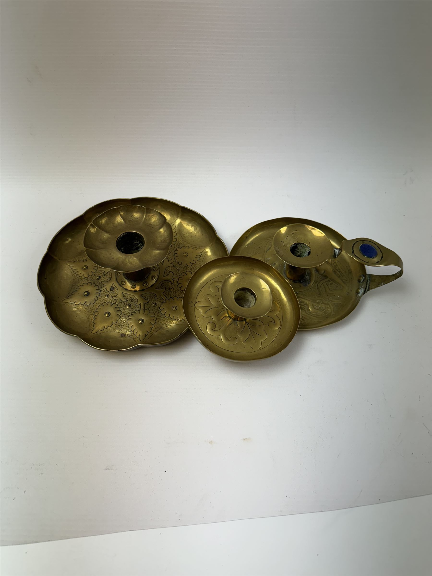 Three Arts & Crafts brass chambersticks/candlesticks, to include a small Keswick School of Industrial Arts example, with repousse leaf decoration, stamped KSIA, a chamberstick with Art Nouveau style decoration to drip pan and blue enamel roundel inset into the loop handle and a larger candlestick, with shaped rim and sconce, repousse decoration and lightly hammered finish, largest D18cm