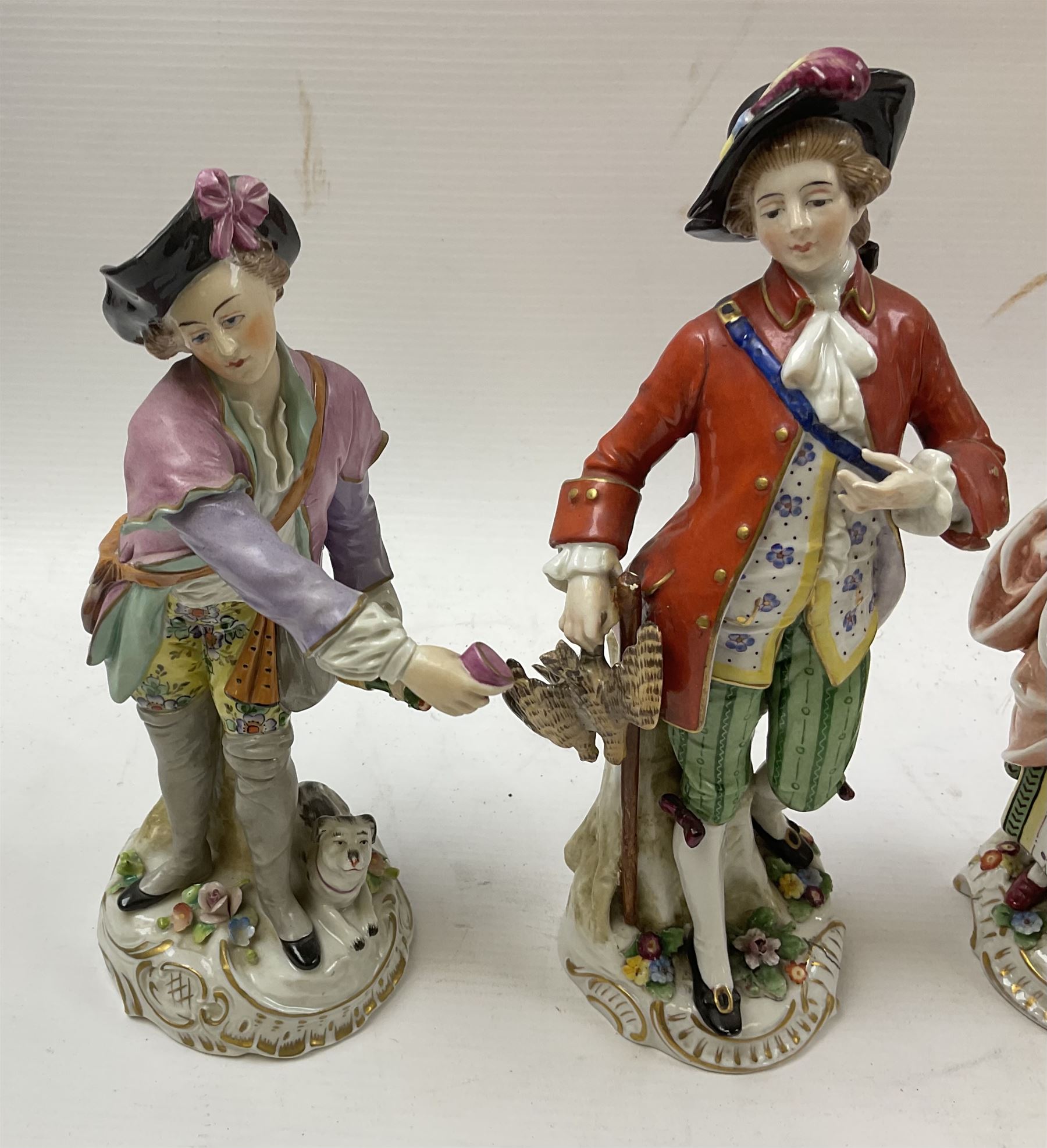 Pair of Naples figures, she holding bird cage with bird perched upon her arm, he holding game, each approximately H23cm, together with a Sitzendorf figure modelled as a figure with wine glass and bottle, with dog laid at feet, H22cm