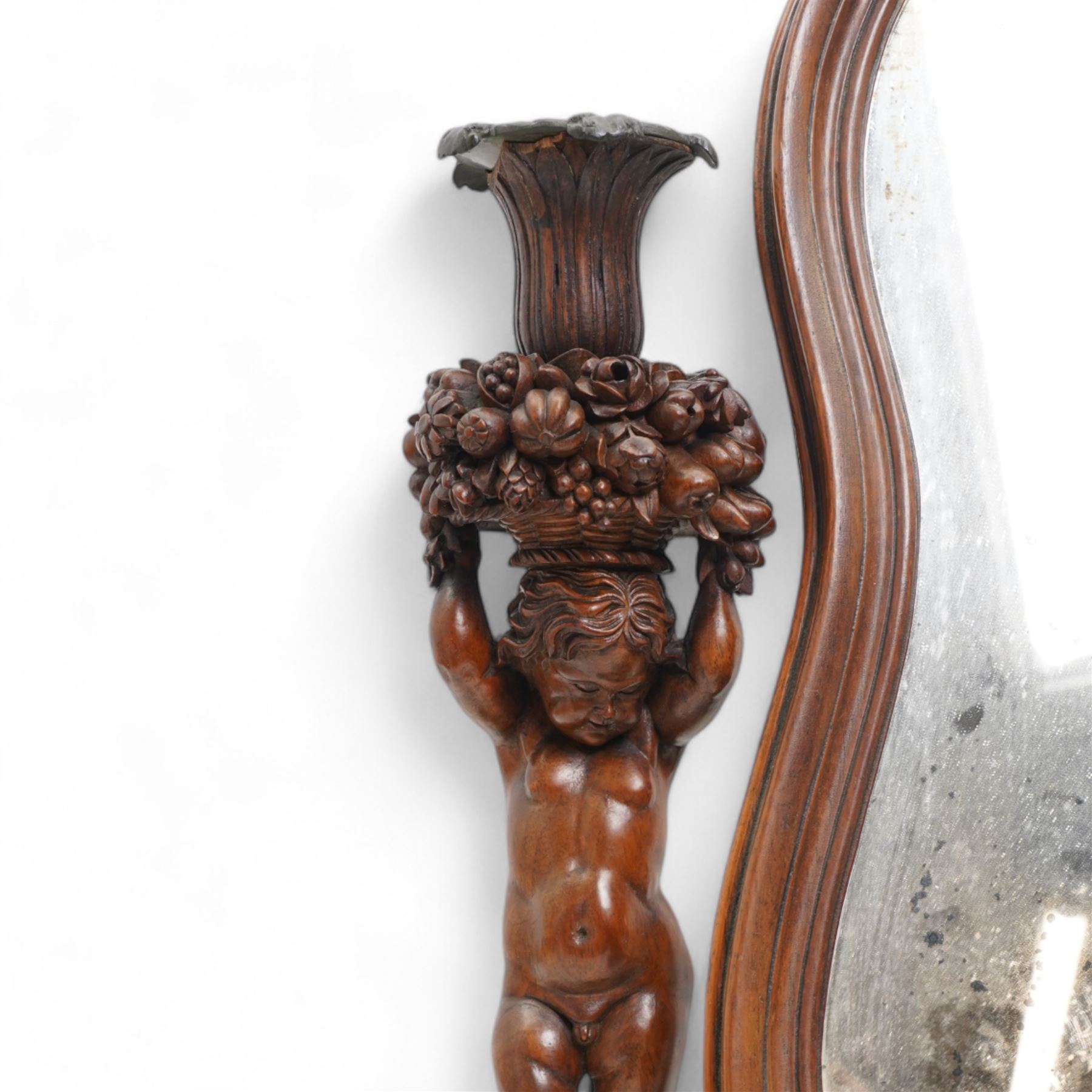 Late 19th century Northern European carved walnut dressing table mirror, the moulded arch top frame carved with flower heads and foliage, flanked by two putto holding baskets of flowers and fruit with candle sconces, on shaped moulded base, plain mirror plate 