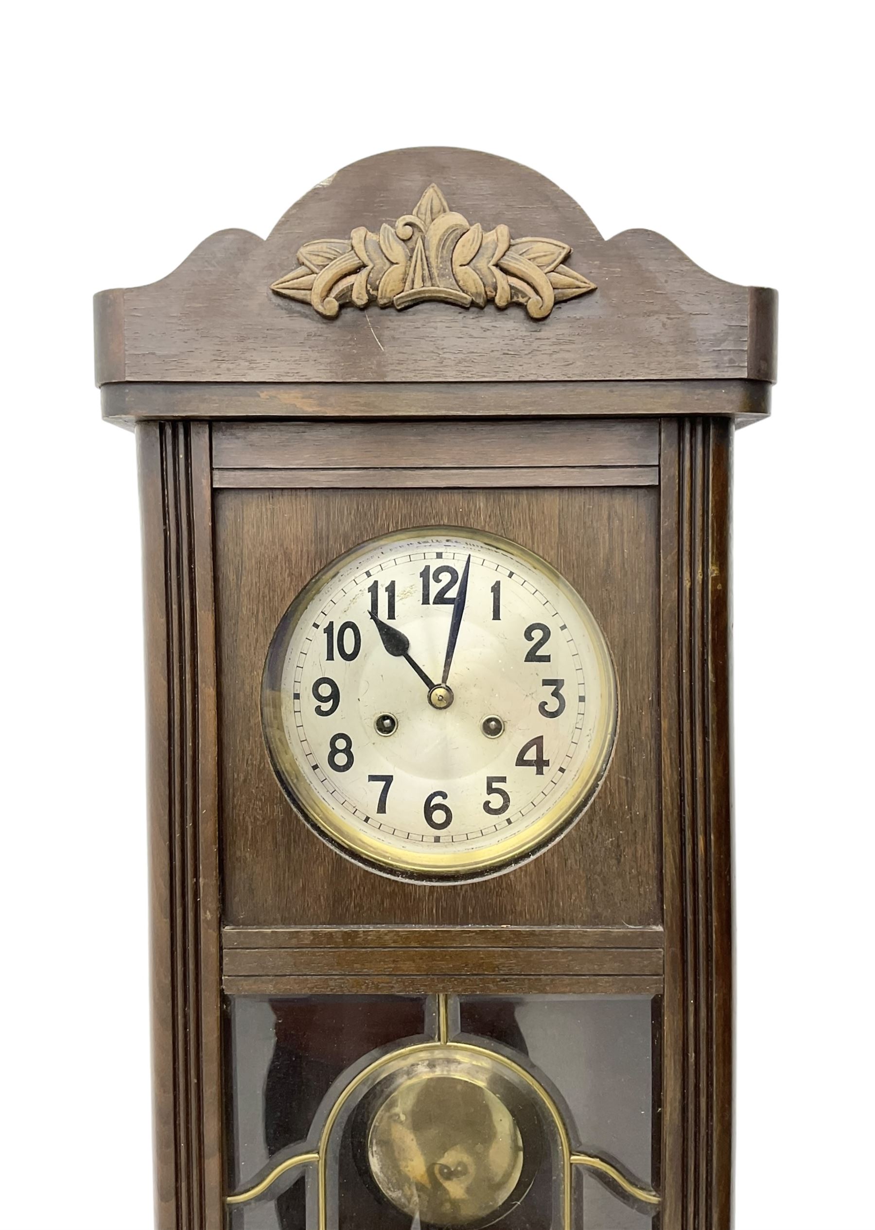 Oak cased 1930s 8-day German wall clock - with a crested top, glazed door and visible pendulum, silvered dial with Arabic numerals and spade hands, two train movement striking the hours on a gong. With pendulum and key.