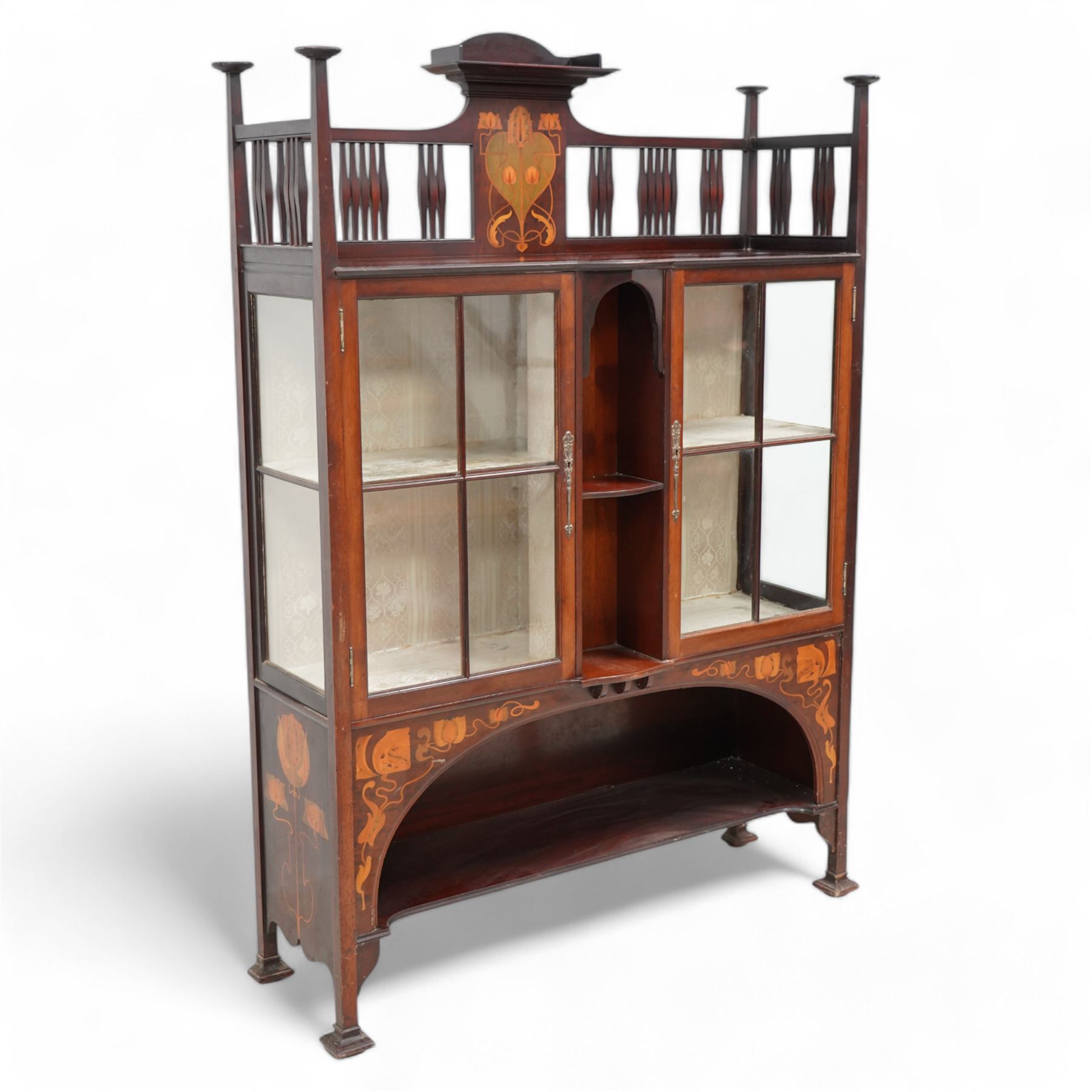 Shapland & Petter; Barnstaple (1854-1999) - Art Nouveau circa. 1900s mahogany display cabinet, raised shelf over panel inlaid with heart and stylised tulips with curled leaves, flanked by balustrade, enclosed by two astragal glazed doors, the lined interior fitted with two shelves, recessed central upright fitted with single shelf, arched lower section fitted with undertier, the spandrels inlaid with further stylised flower heads and trailing branches, on style supports terminating to moulded pad feet, the locks stamped 'S&B, B', retailed by 'Denby & Spinks, Leeds' plaque verso