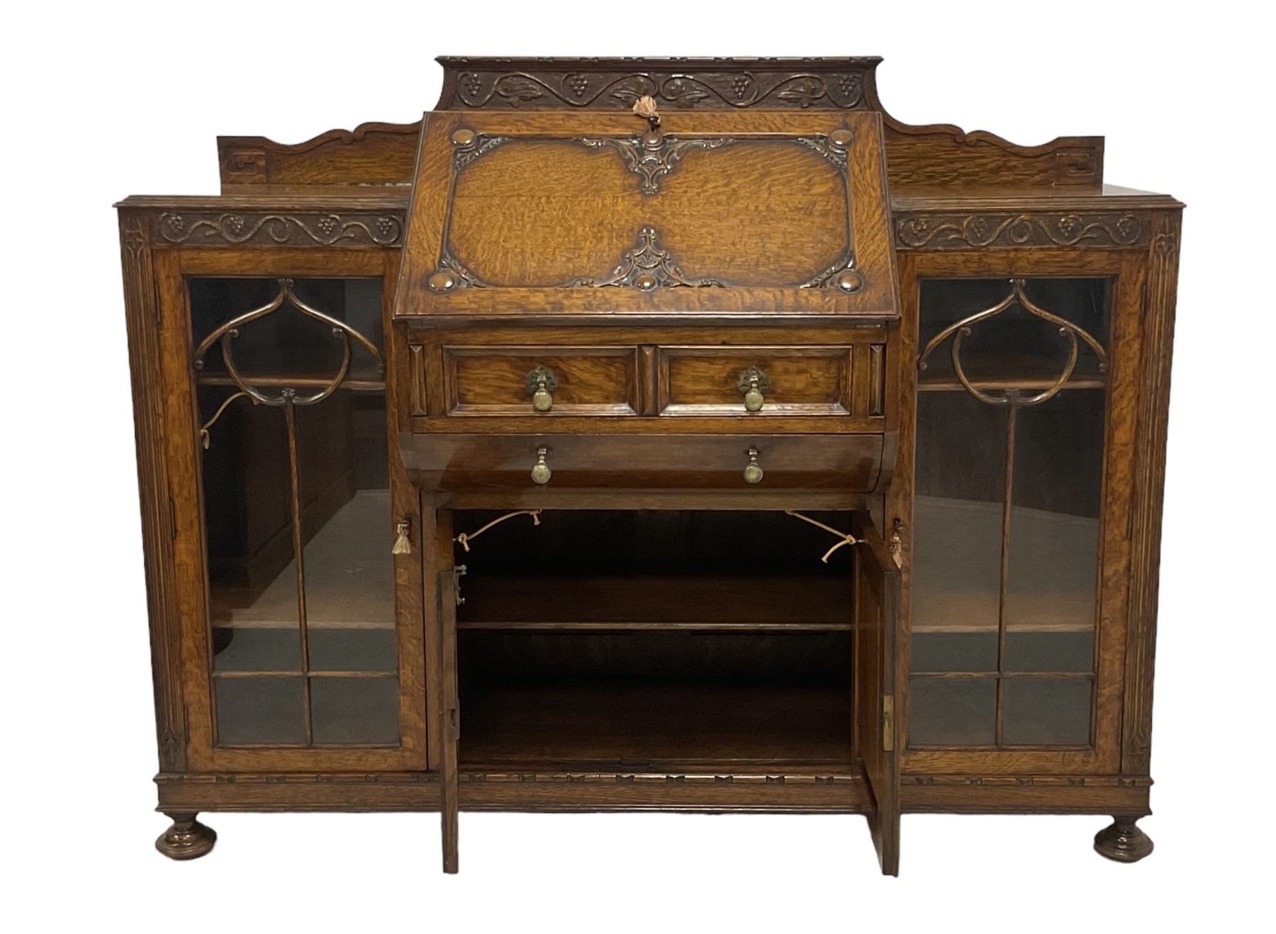 Early 20th century heavily carved oak bookcase bureau, raised back with grape-vine carved frieze, over fall-front enclosing fitted interior, above two short drawers and curved frieze drawer, flanked by astragal glazed and panelled cupboards, on turned feet