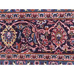 Large Kashan crimson ground carpet, central indigo medallion with stylised floral motifs, flanked by matching spandrels, bordered by multiple bands featuring repeating foliate and floral designs throughout