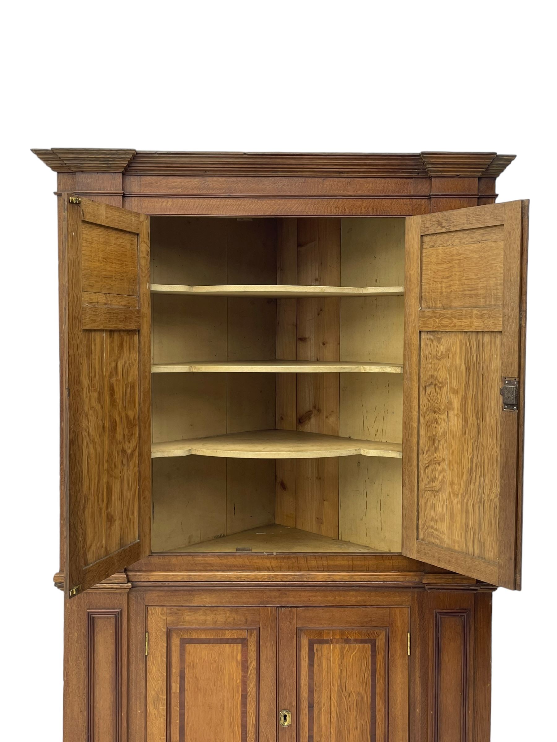 George III and later oak and mahogany banded corner cupboard, moulded cornice over four panelled doors, the door panels inlaid with mahogany bands, projecting square pilasters throughout with applied reeded mouldings, the painted interior fitted with shaped shelves, on moulded plinth base 