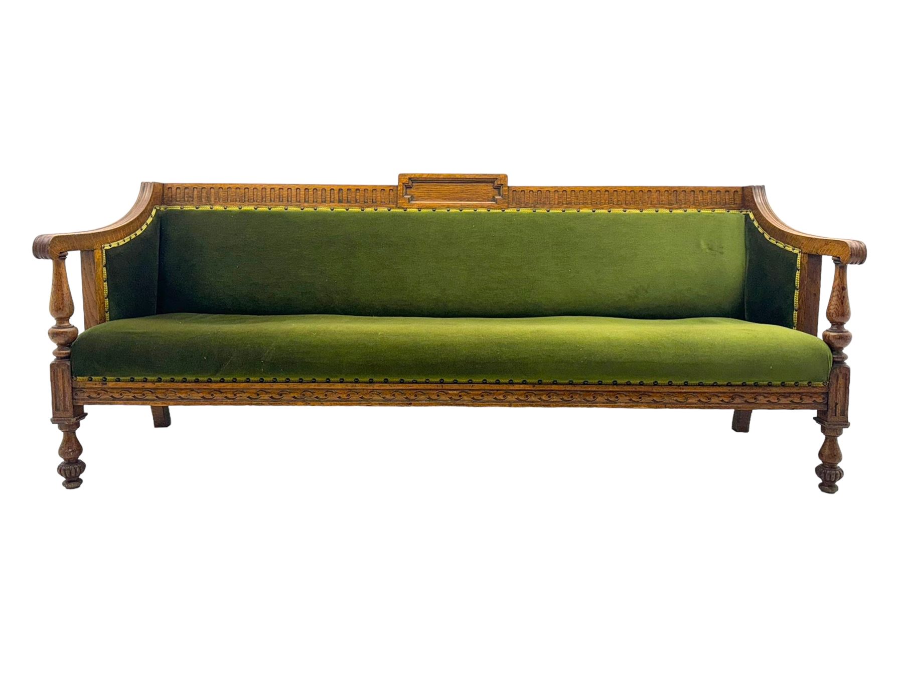 Late Victorian walnut framed sofa, rectangular backrest with carved detailing and central motif, upholstered in olive green fabric with brass studded trim and patterned front rail, scrolled fluted arms supported by turned uprights terminating in bun feet