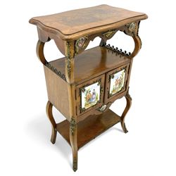 Mid-to-late 20th century French figured walnut three-tier bedside stand, shaped moulded top inlaid with figured book-matched veneers and crossbanding, double cupboard below enclosed by two doors mounted by painted porcelain panels depicting courting scenes within landscapes, on cabriole supports united by undertier, decorated with ornate cast metal cartouche mounts 