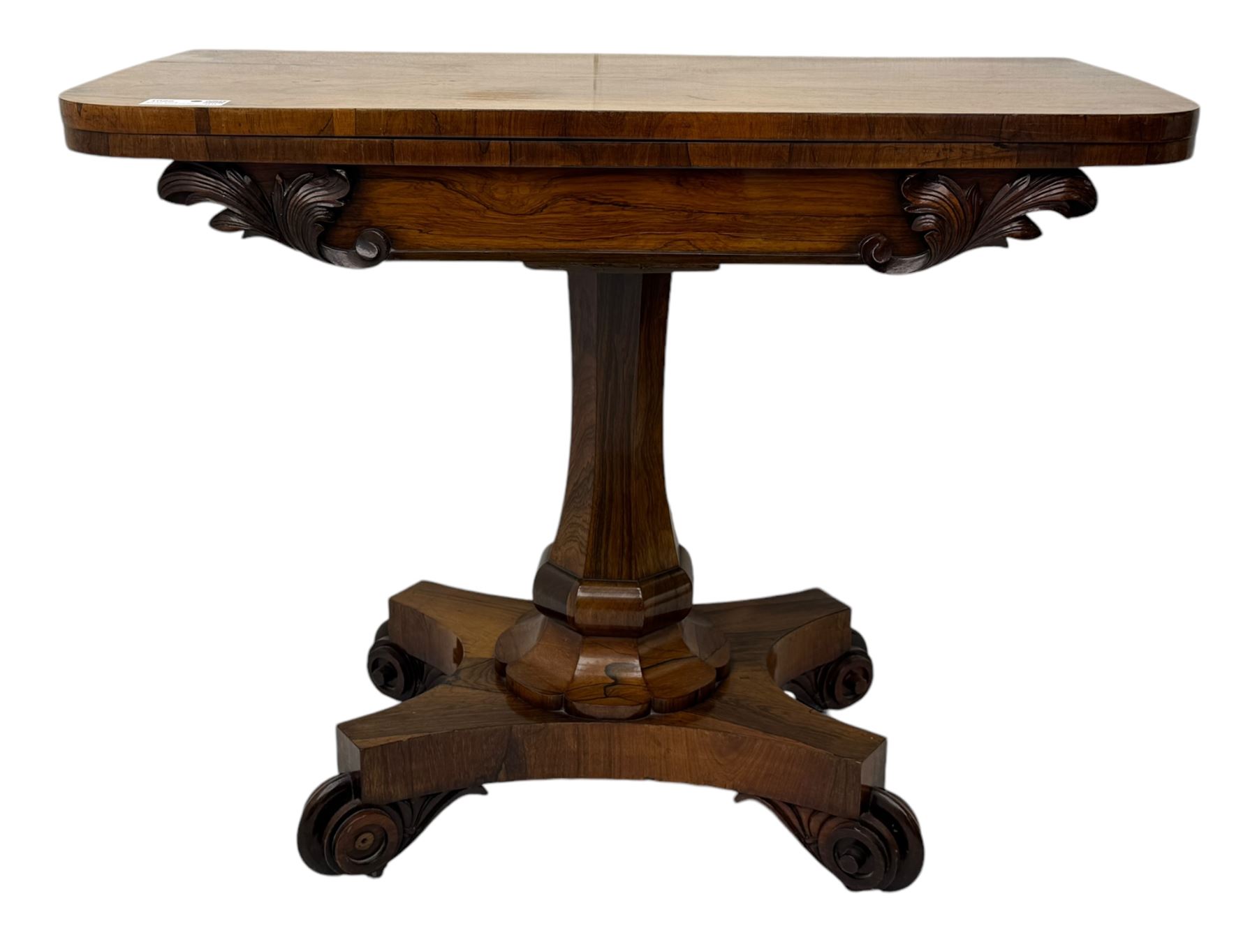 Early Victorian rosewood card table, rectangular foldover swivel top with rounded corners, red baize-lined surface, frieze adorned with carved foliate motifs, supported by a central octagonal waisted column on shaped quatrefoil base, terminating in scrolled feet with concealed metal castors