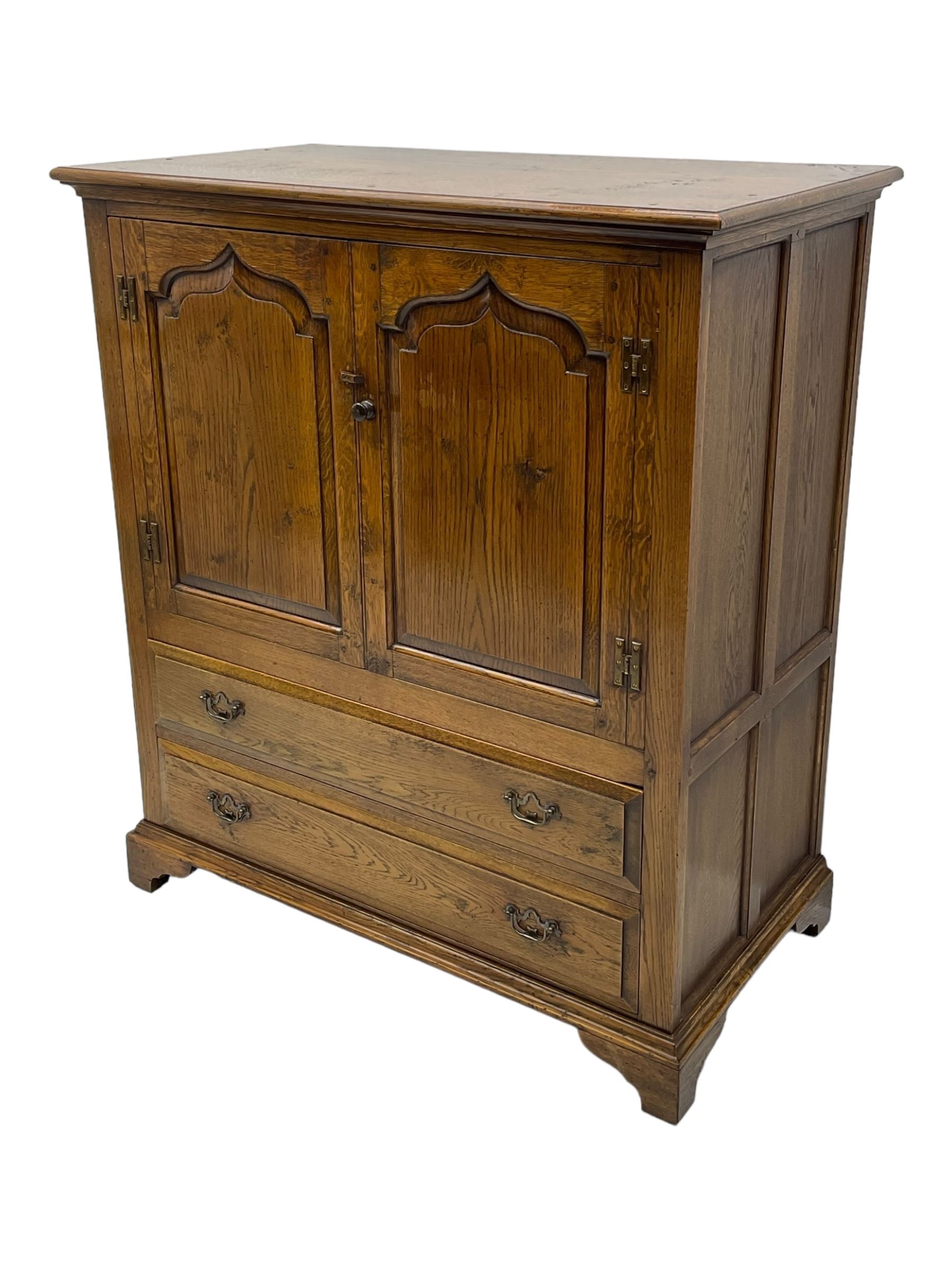 Oak drinks or media cabinet, projecting moulded cornice over two Gothic arch panelled doors on sliding tracks enclosing open storage, two drawers with brass drop handles, on bracket supports