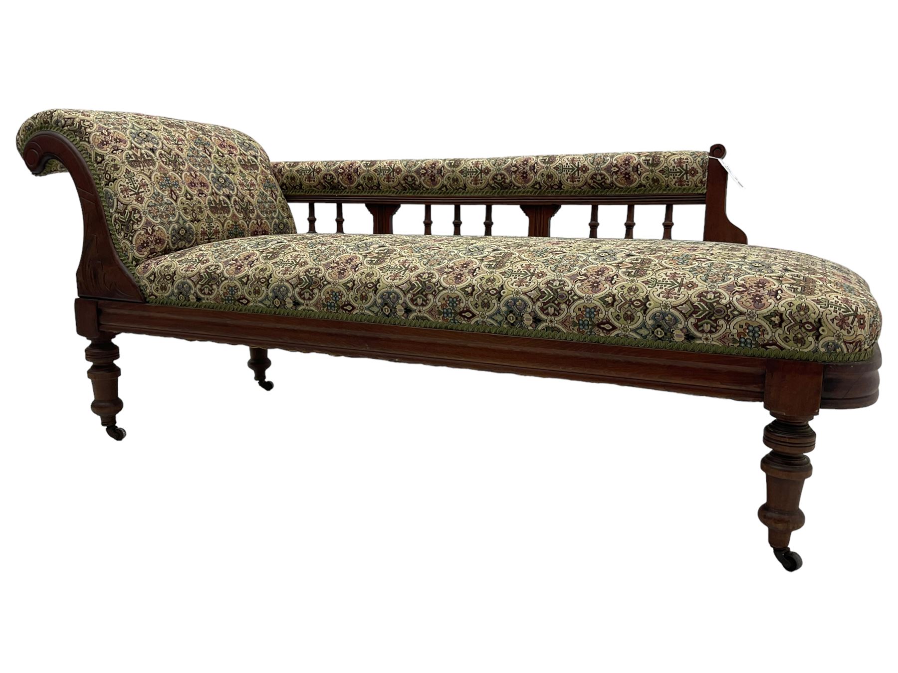 Late 19th century walnut framed chaise longue, upholstered in floral pattern fabric, rolled back rest and turned balustrade back, on turned feet with brass and ceramic castors 