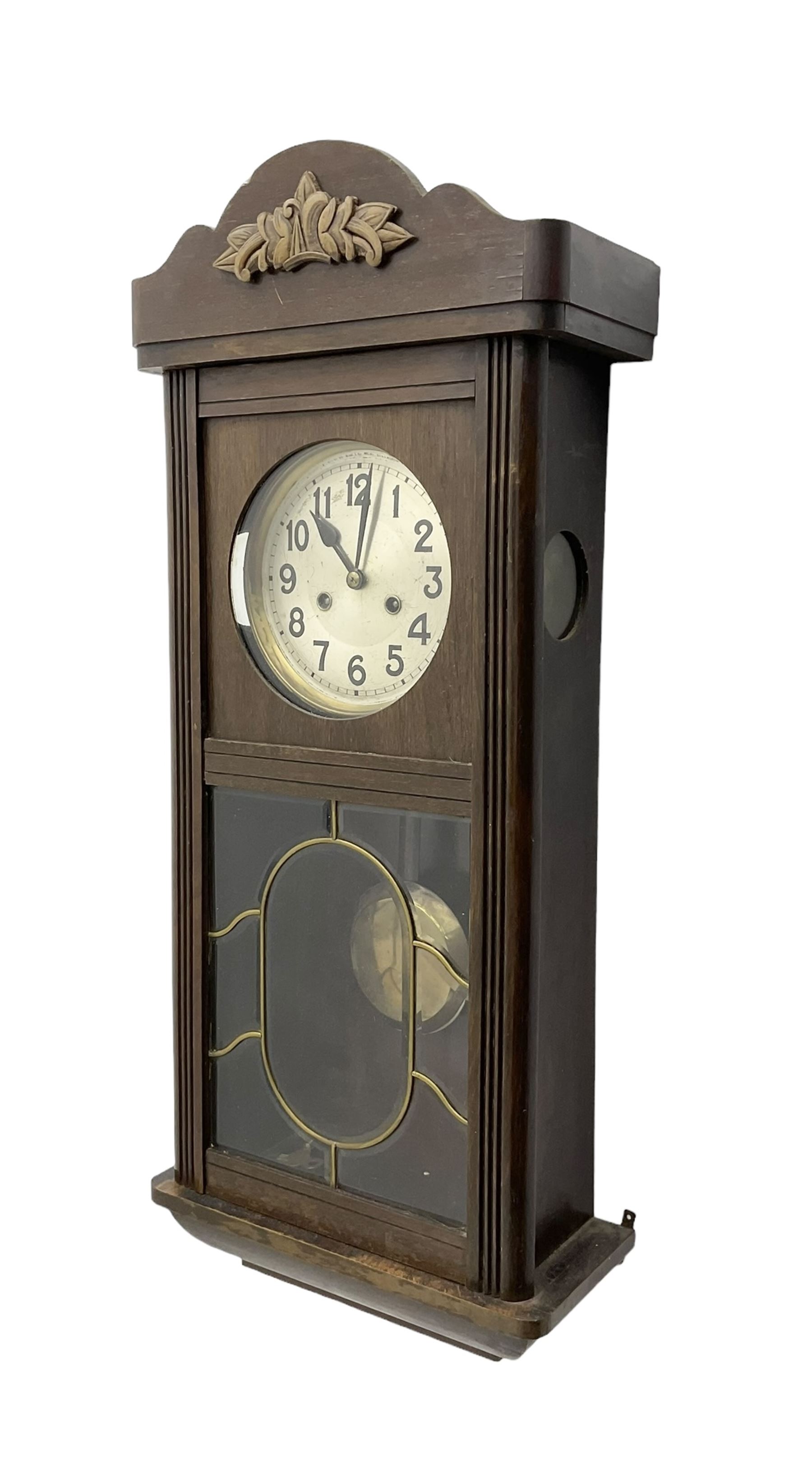 Oak cased 1930s 8-day German wall clock - with a crested top, glazed door and visible pendulum, silvered dial with Arabic numerals and spade hands, two train movement striking the hours on a gong. With pendulum and key.