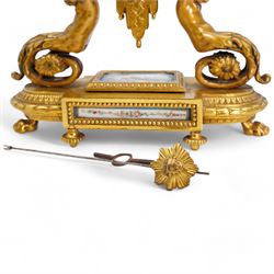 French-mid 19th century 8-day gilt and porcelain mantle clock with a break front plinth raised on paw feet, with recessed rectangular porcelain panels depicting two lovers and floral decoration, drum cased movement with a porcelain dial supported on two matching scroll supports, surmounted with a tapered porcelain urn and gilt finial, dial with cartouche Roman numerals on a blue ground and a depiction of cupid to the dial centre, twin train rack striking movement, striking the hours and half hours on a bell. With Pendulum and key.