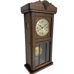 1930s wall clock in an oak case with a crested pediment and glazed door with visible pendulum, silvered dial with Roman numerals and steel spade hands, eight day spring driven movement striking the hours and half hours on a gong. With pendulum and key.