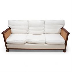 Early 20th century mahogany framed bergère three seat sofa, upholstered back and seat flanked by rolled double-caned arms with carved scale decoration, floral and ribbon-twist carved lower edge, over ball-and-claw feet