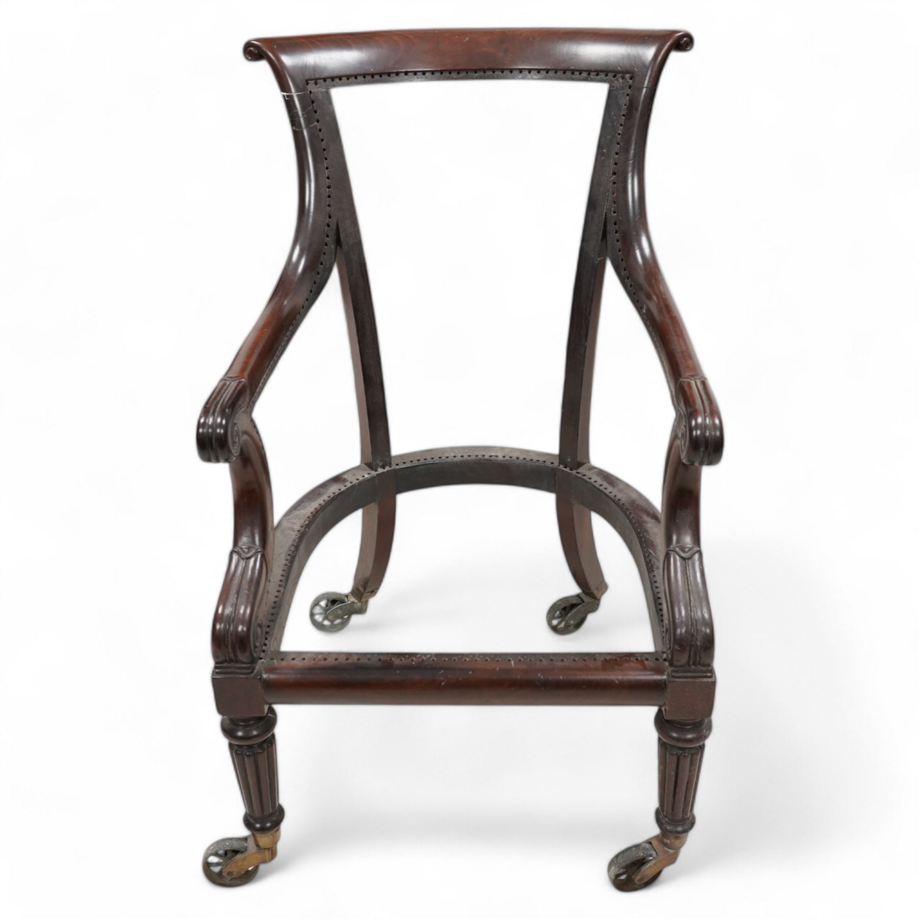 Possibly Gillows - Regency mahogany library chair frame, curved and rolled cresting rail over sweeping arms with lappet and scroll carved terminals, on turned and reed carved supports, brass cups and spoked wheel castors 