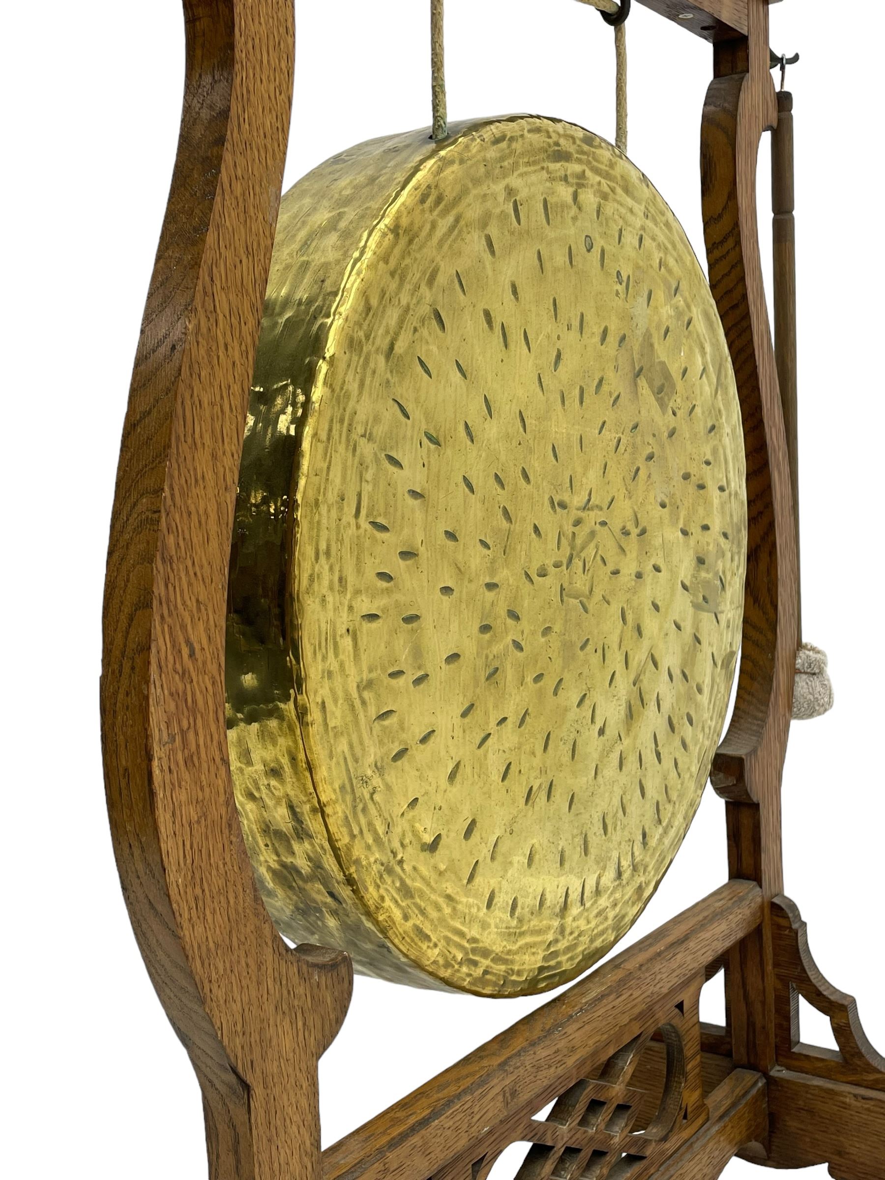 Early 20th century oak gong stand with gong and hammer, shaped cresting rail over moulded upper edge on shaped supports, hanging brass drum gong over fretwork middle rail, on raised platforms terminating to splayed feet