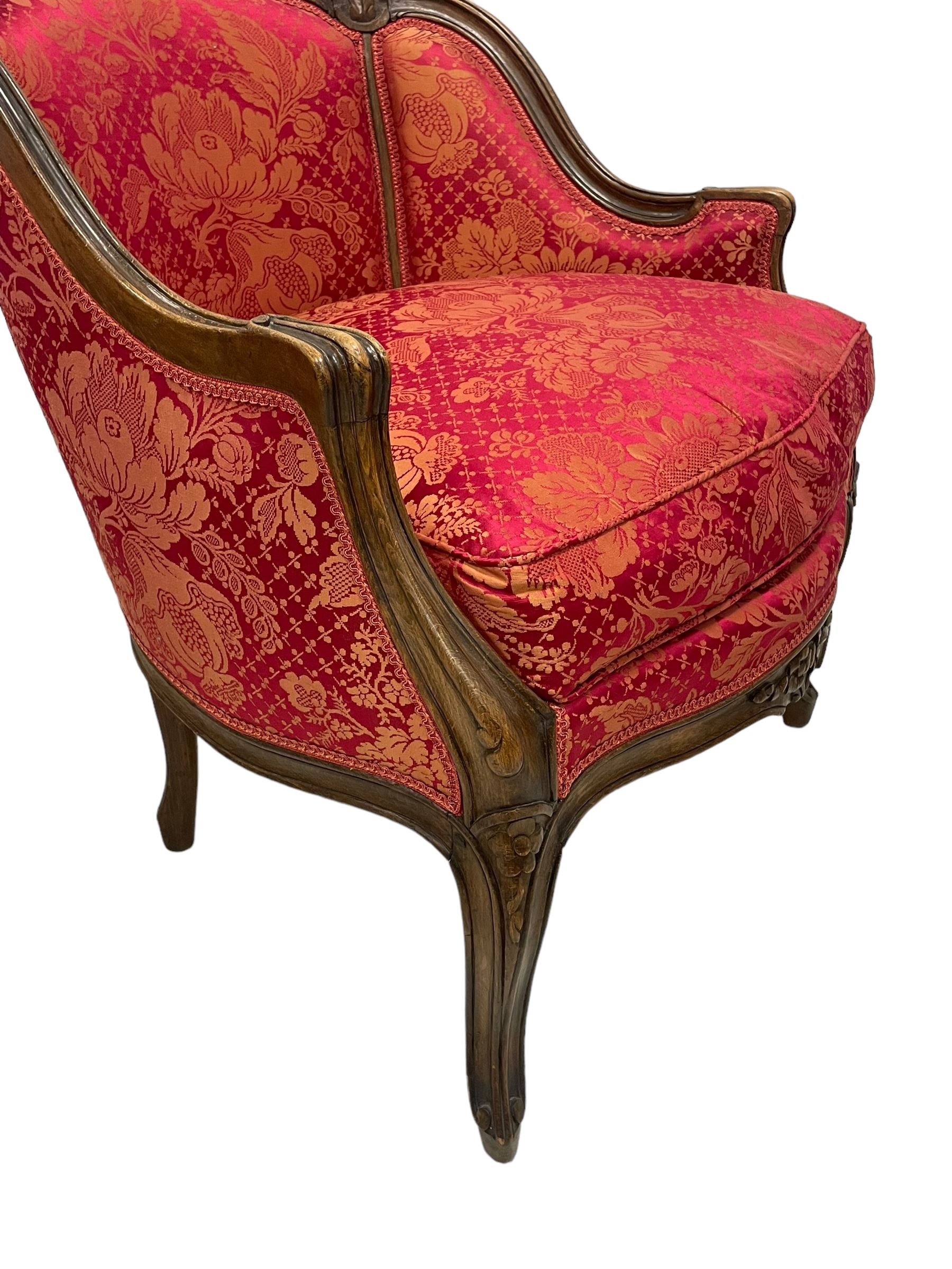 Late 20th century French design stained beech framed armchair and matching stool - the armchair with shaped and moulded frame carved with flower heads and foliage, upholstered in silk red and gold ground fabric with floral pattern, on scroll carved cabriole feet decorated with flower head carvings (W73cm, H82cm, D75cm); the footstool of concaved rectangular form with moulded frame carved with flower heads, upholstered loose cushion, on cabriole feet (70cm x 64cm, H48cm)