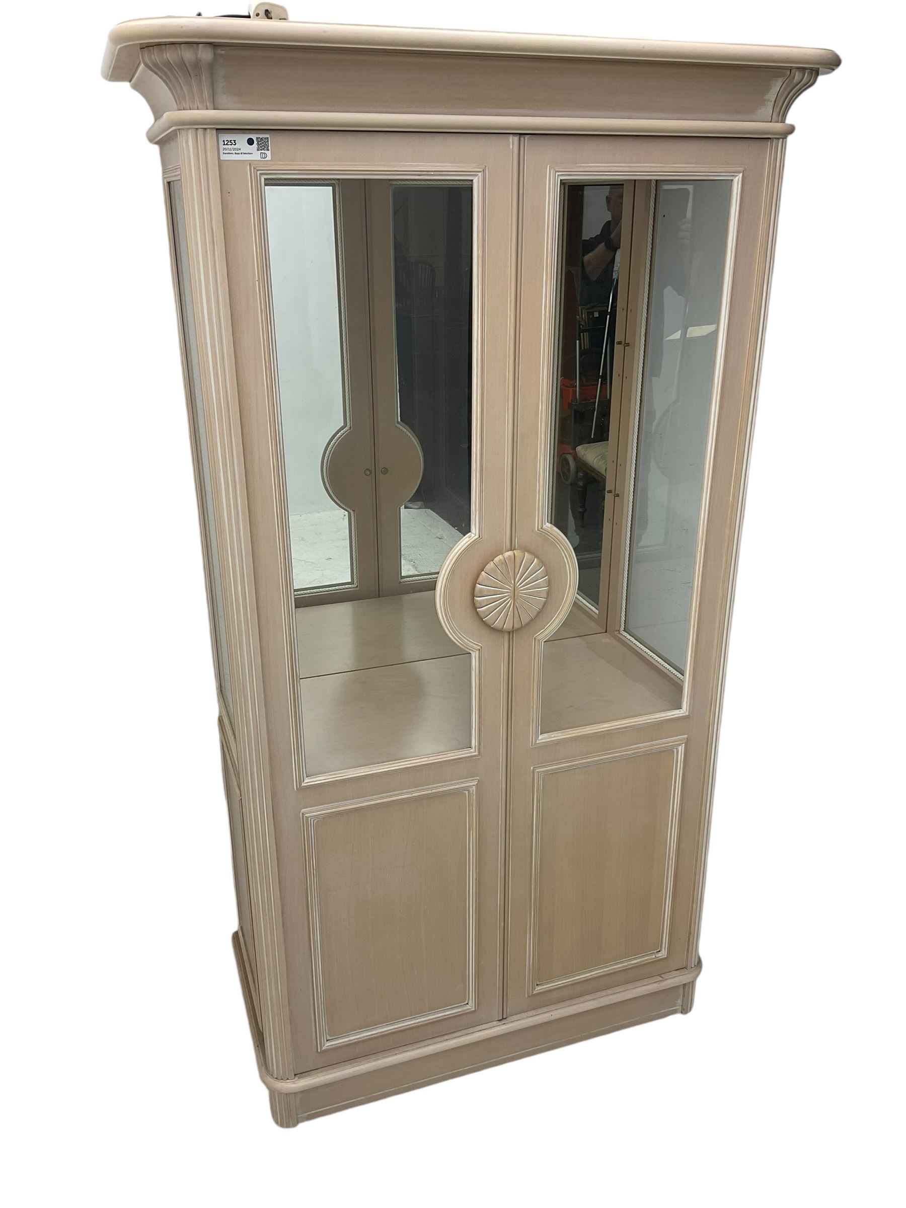 Contemporary display cabinet, moulded cornice above glazed panelled doors with central carved sunburst handles, mirrored back interior with lighting, lower enclosed storage compartment, raised on plinth base