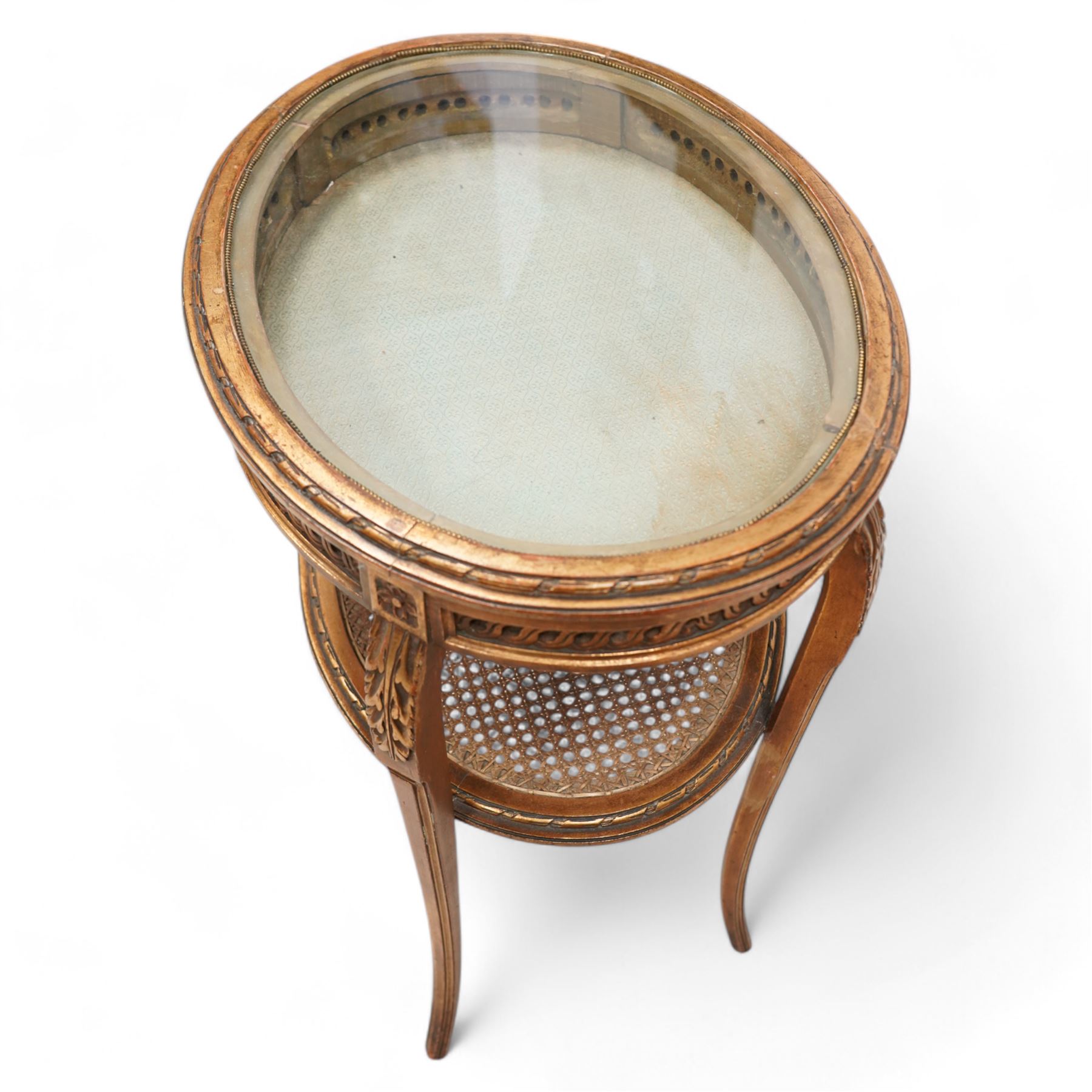 Late 19th century gilt wood bijouterie table, oval form, lift-up glazed lid with ribbon moulded edge, pierced and guilloche carved frieze rails, acanthus carved and moulded cabriole supports united by canework undertier  