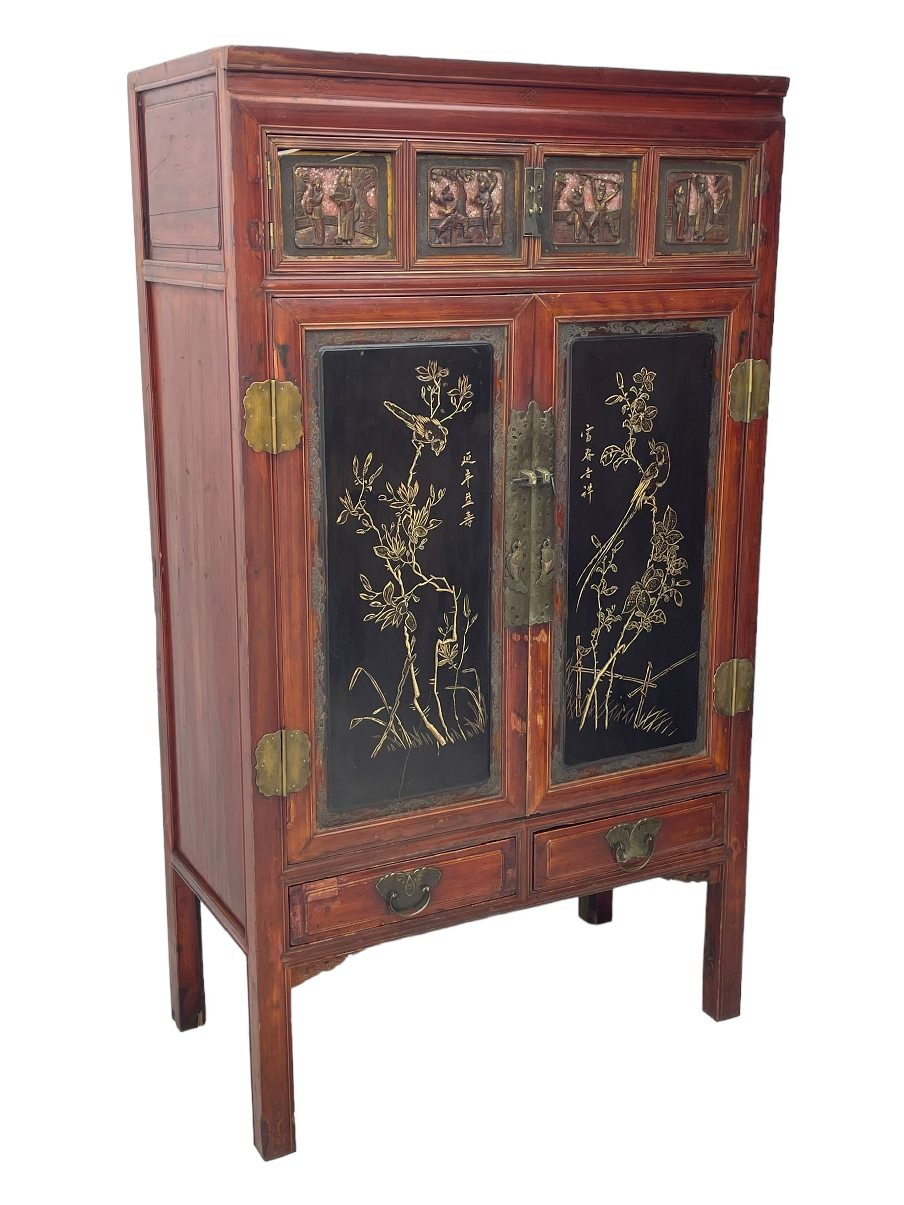 Late 19th century Chinese Qing dynasty red and black lacquered wood cabinet, Fujianese province, upper cupboard enclosed by two doors with relief carved and gilt panels depicting figures behind glass, the large cupboard enclosed by two panelled doors carved with trailing foliate branches and birds, inscribed with Chinese characters, to the left ‘Prolong life’ and to the right ‘Wealth and good fortune’, fitted with two drawers, square supports with carved brackets 