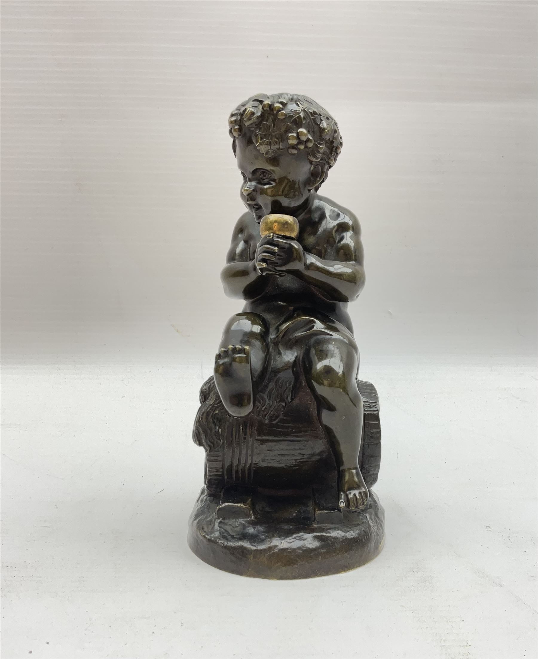 Bronzed metal model of a putti seated upon a barrel with goblet in hands, and fruiting vines adorning their head, H17.5cm