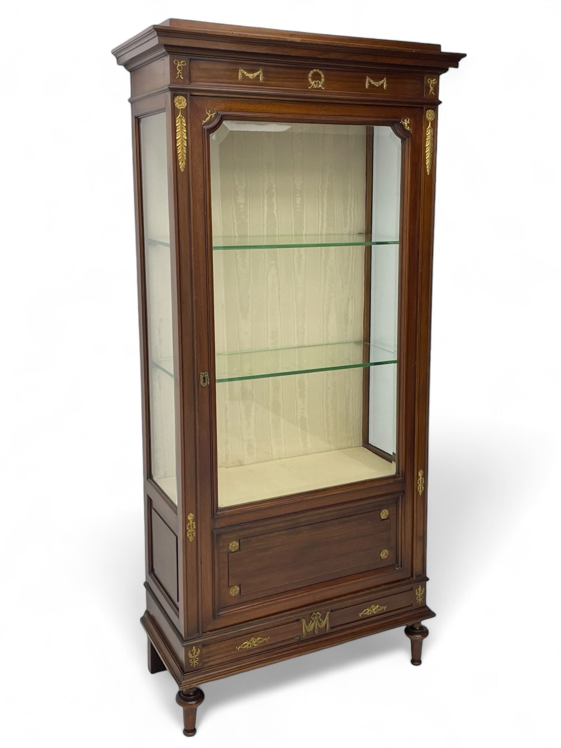 Early 20th century Louis XVI design mahogany vitrine display cabinet, projecting stepped cornice over recessed frieze decorated with gilt metal mounts of festoons and a bellflower wreath, fabric lined interior with two glass shelves, enclosed by bevel glazed door with lower panel, on two turned front feet 