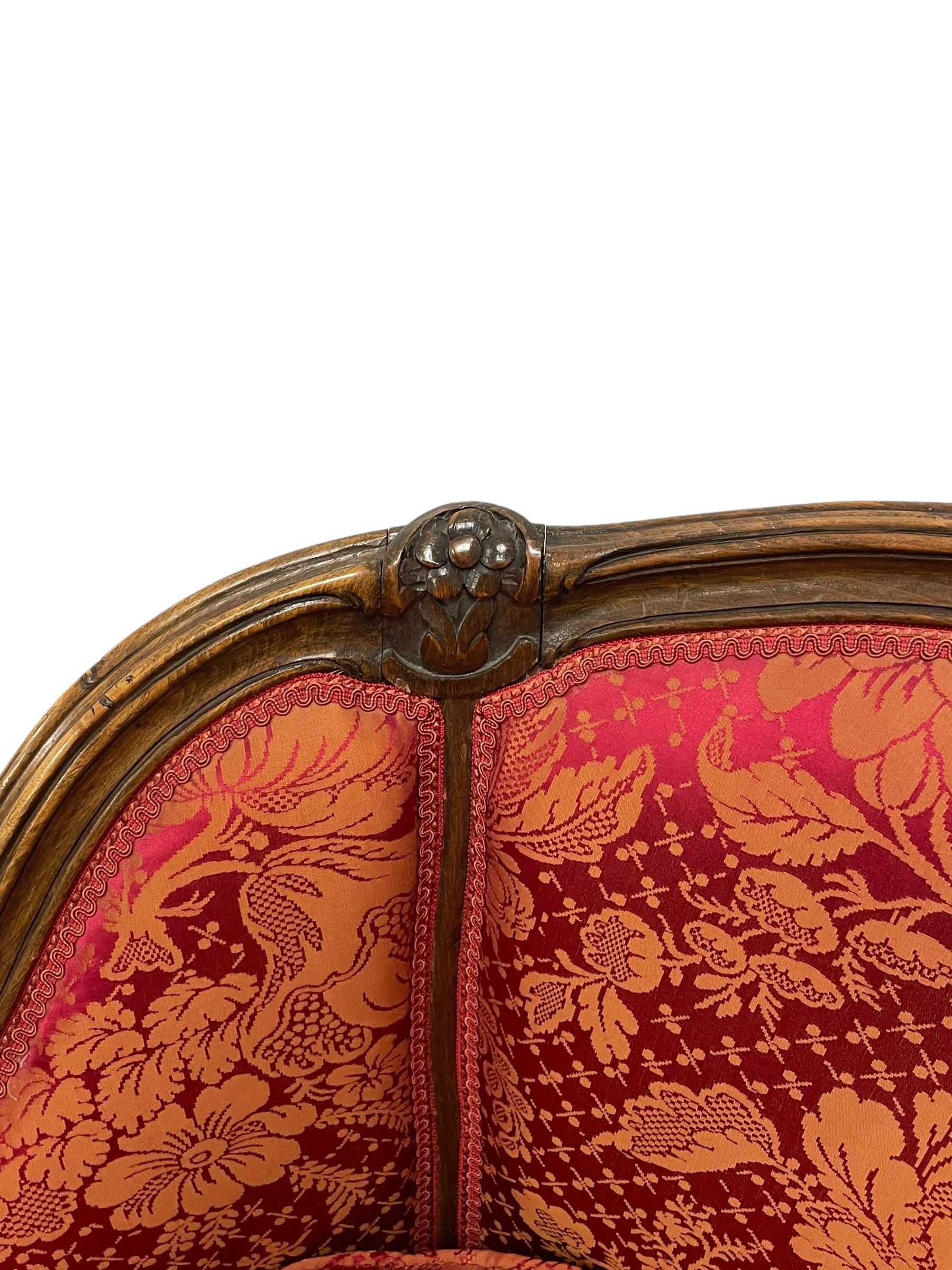 Late 20th century French design stained beech framed armchair and matching stool - the armchair with shaped and moulded frame carved with flower heads and foliage, upholstered in silk red and gold ground fabric with floral pattern, on scroll carved cabriole feet decorated with flower head carvings (W73cm, H82cm, D75cm); the footstool of concaved rectangular form with moulded frame carved with flower heads, upholstered loose cushion, on cabriole feet (70cm x 64cm, H48cm)