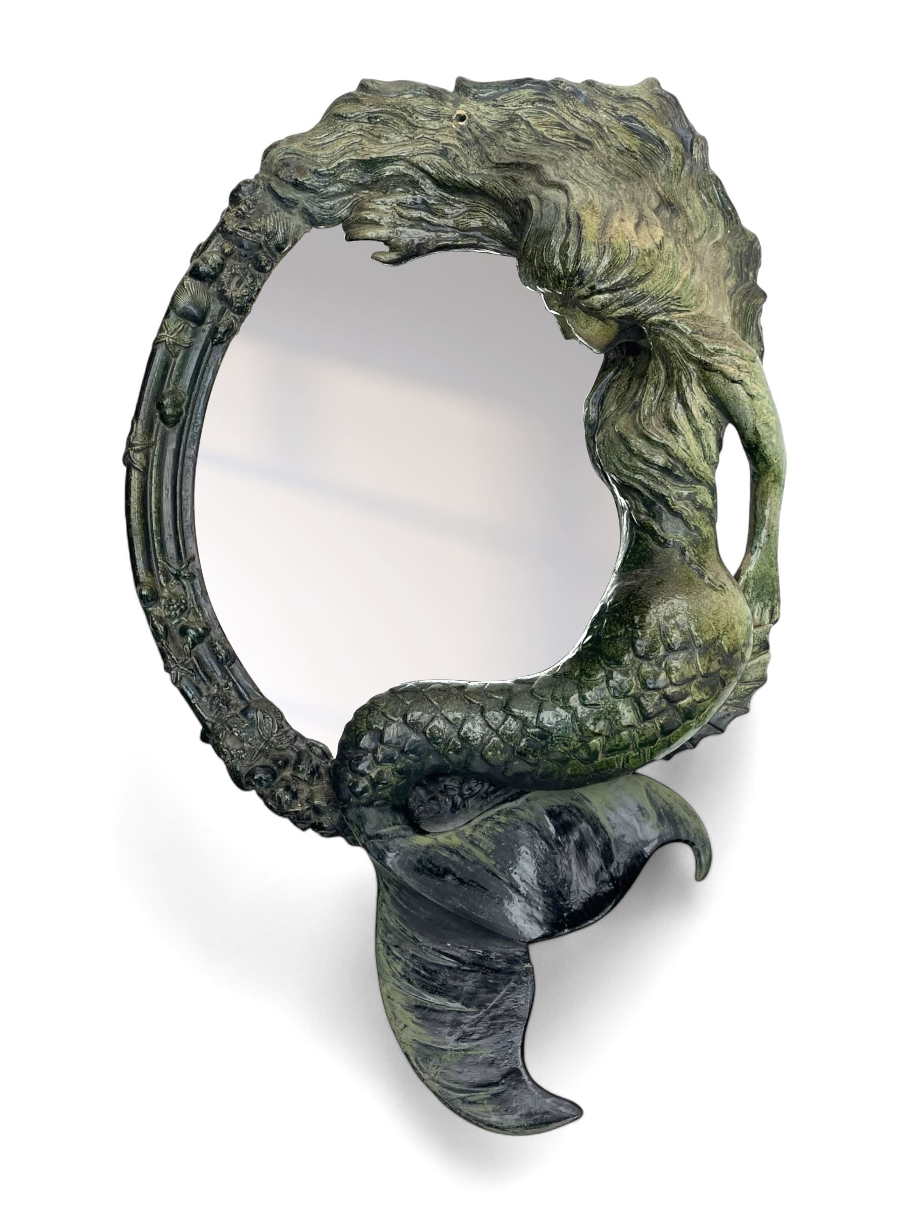 French Art Nouveau style wall mirror, the oval mirror plate within a stepped frame adorned with sea shells and mounted with the figure of a mermaid with flowing hair, her arm reaching across the mirror plate, the whole with green glazed finish, overall approximately H100cm W60cm