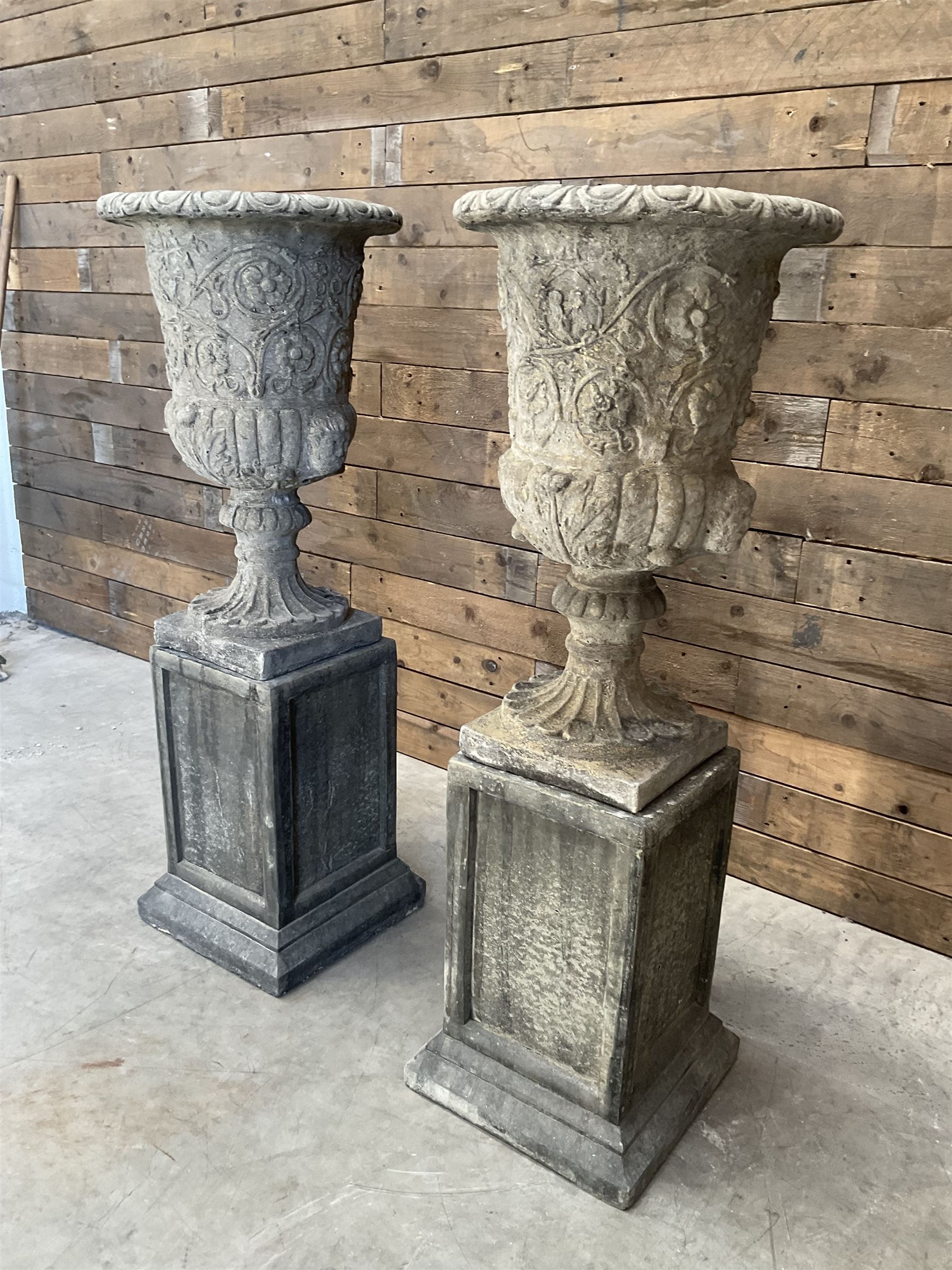 Pair of Georgian design cast stone garden urns, egg and dart rim with raised floral motifs, pedestal base terminating on square plinth, raised on square block plinth