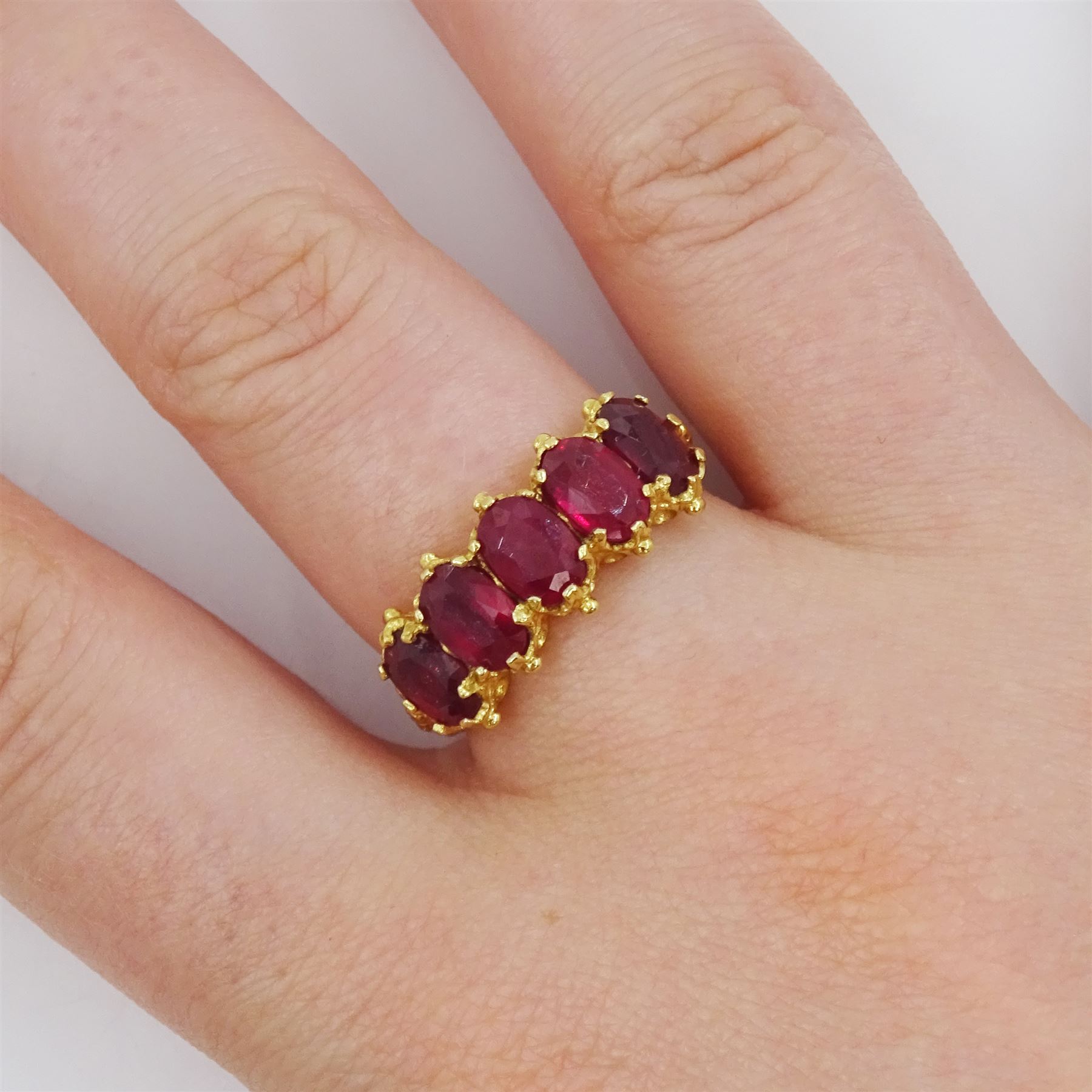 18ct gold five stone oval cut glass-filled ruby ring, with scroll design shoulders, London 2005