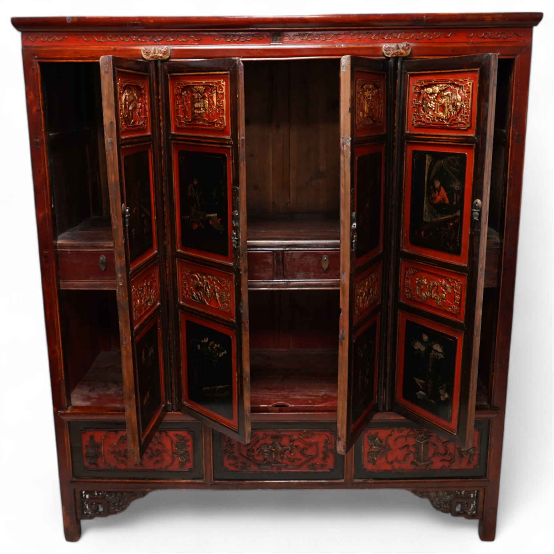 19th century Chinese red and black lacquered hardwood cabinet fitted with four panelled doors, the frieze decorated with scrolling gilt patterns over gold bat mounts surmounting the hinges, the black panels depicting traditional figural and floral scenes, the carved parcel-gilt panels with pagoda, pastoral and war scenes, the doors fitted with metal fish and pagoda handles, enclosing a single shelf and four internal drawers, the base compartment decorated with carved precious objects, raised on moulded square supports with pierced scrolling brackets