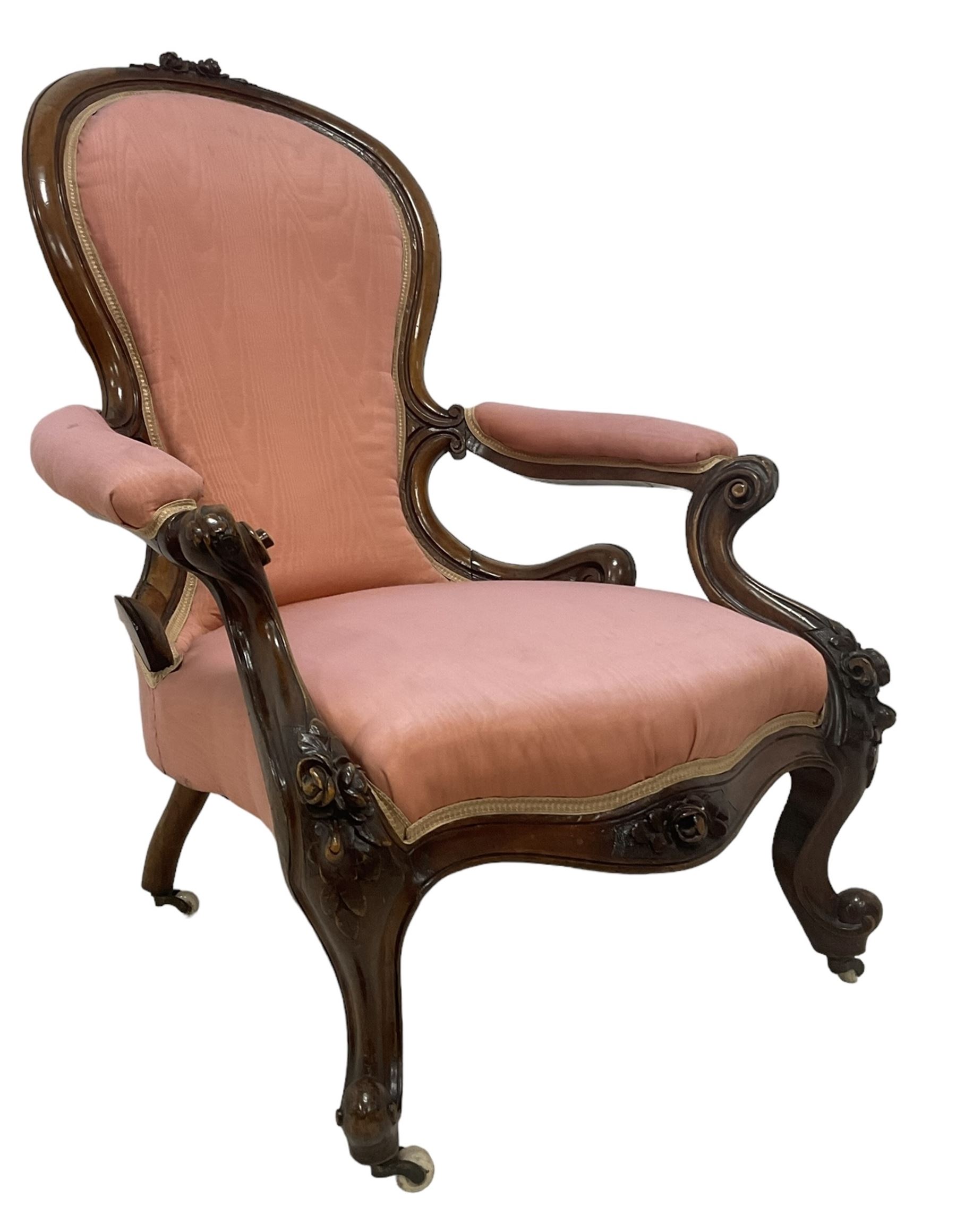 Victorian mahogany framed open armchair, spoon-back and sprung seat upholstered in pink fabric, scrolled arm terminals over serpentine fronted seat decorated with moulded carved floral motifs, cabriole supports with ceramic castors