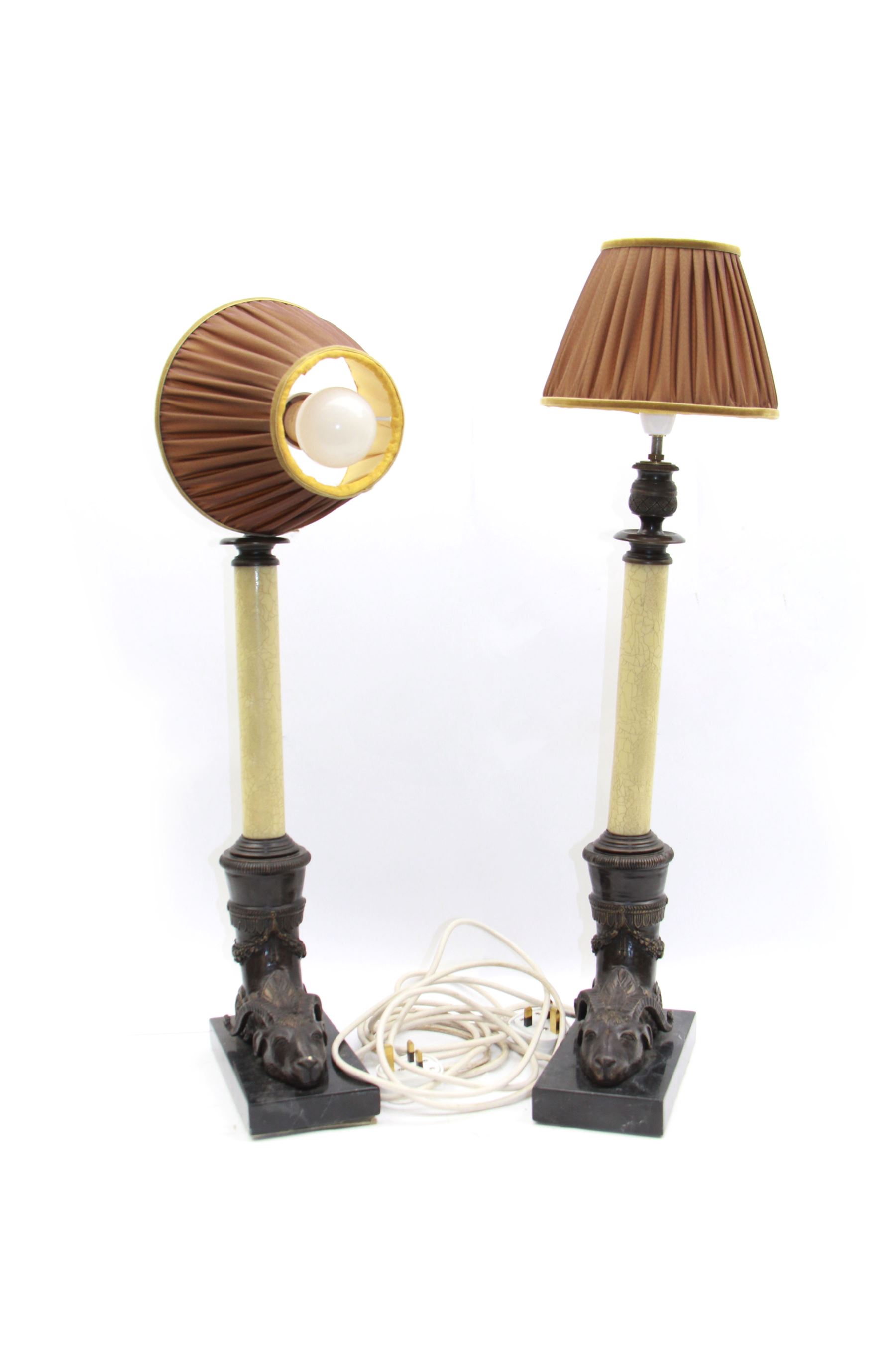 Pair of bronze effect brass table lamps, modelled as cornucopia issuing from rams heads, leading to a crackle effect column supporting pleated fabric shades, the whole upon black stone plinths, overall H67cm 