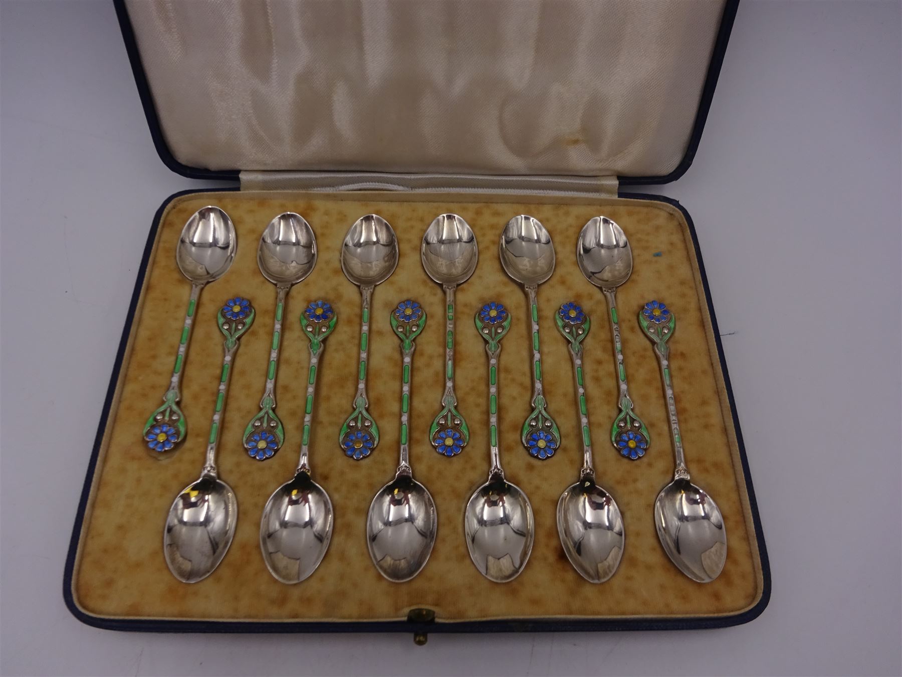 Set of twelve silver coffee spoons, each with enamelled blue flower to terminal and green and white enamel decoration to stem, hallmarked Mappin & Webb Ltd, Birmingham 1934, contained within fitted case