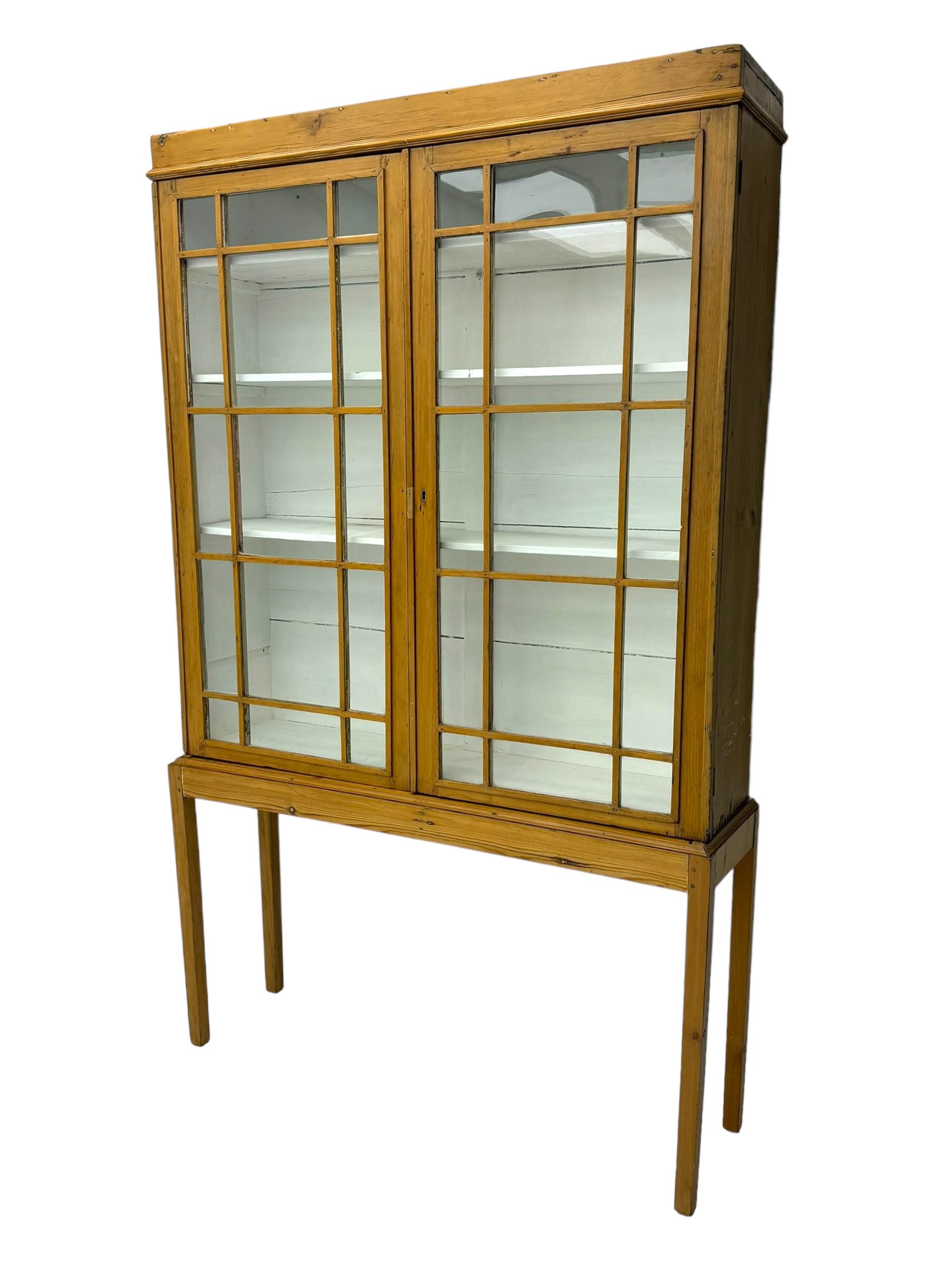 Georgian stripped pine bookcase display cabinet, plain moulded cornice over two glass-panel doors with multiple panes, enclosing white painted interior with three shelves, raised on square supports