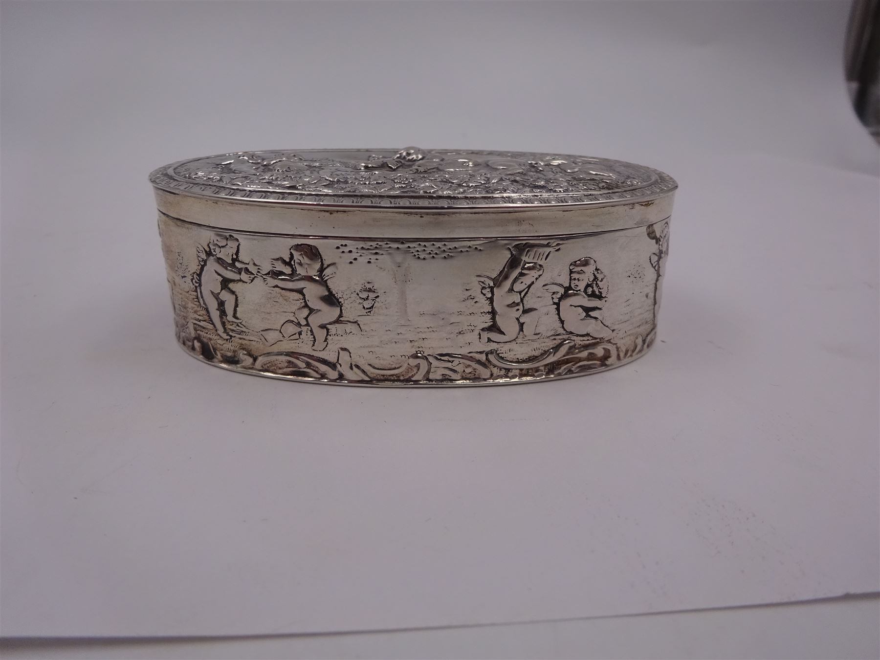 Late 19th century German Hanau silver box, of oval form, embossed throughout with putti in various scenes including riding a dog, playing instruments, pulling a cart and dancing, opening to reveal a gilt interior, with Hanau marks for B Neresheimer & Sohne, W9cm, H3cm