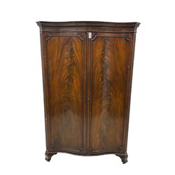 Early 20th century mahogany serpentine double wardrobe, moulded cornice over two figured doors, the interior fitted with hanging rail and shelf, canted and fluted uprights, on bracket feet 