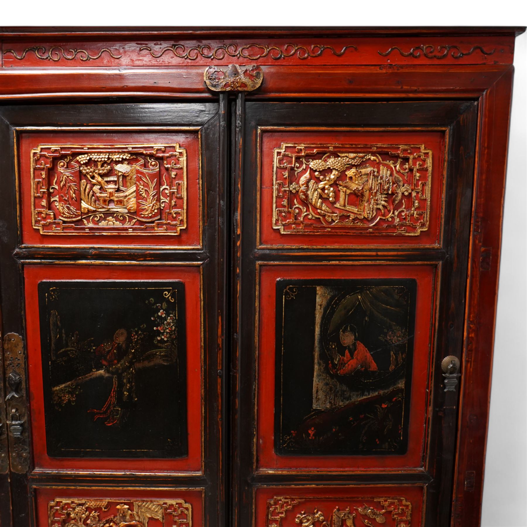 19th century Chinese red and black lacquered hardwood cabinet fitted with four panelled doors, the frieze decorated with scrolling gilt patterns over gold bat mounts surmounting the hinges, the black panels depicting traditional figural and floral scenes, the carved parcel-gilt panels with pagoda, pastoral and war scenes, the doors fitted with metal fish and pagoda handles, enclosing a single shelf and four internal drawers, the base compartment decorated with carved precious objects, raised on moulded square supports with pierced scrolling brackets