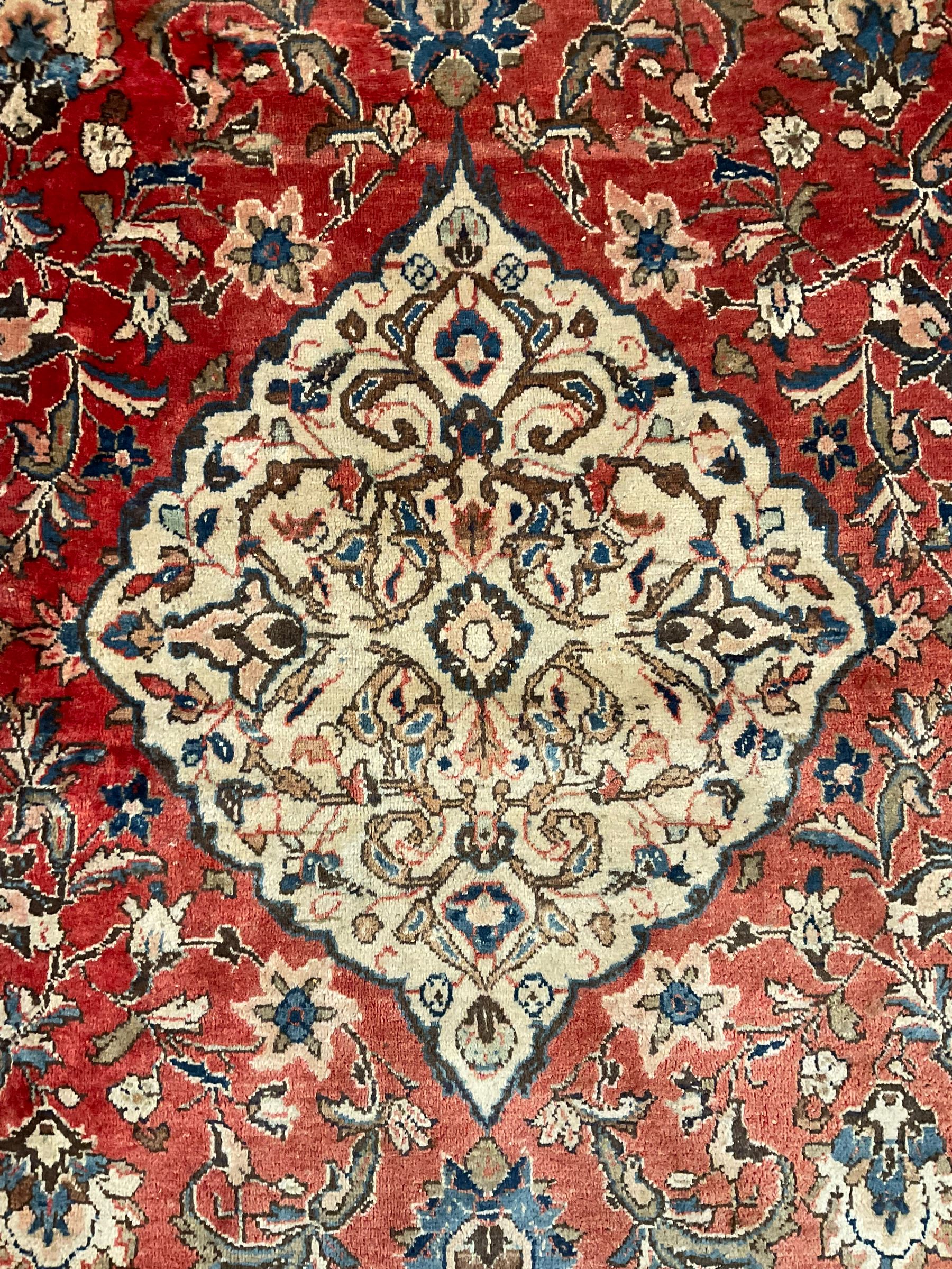 Persian Kashan crimson ground carpet, central shaped pale ground medallion surrounded by interlacing branches and stylised floral motifs, indigo main border with scrolling foliage design, within guard stripes 