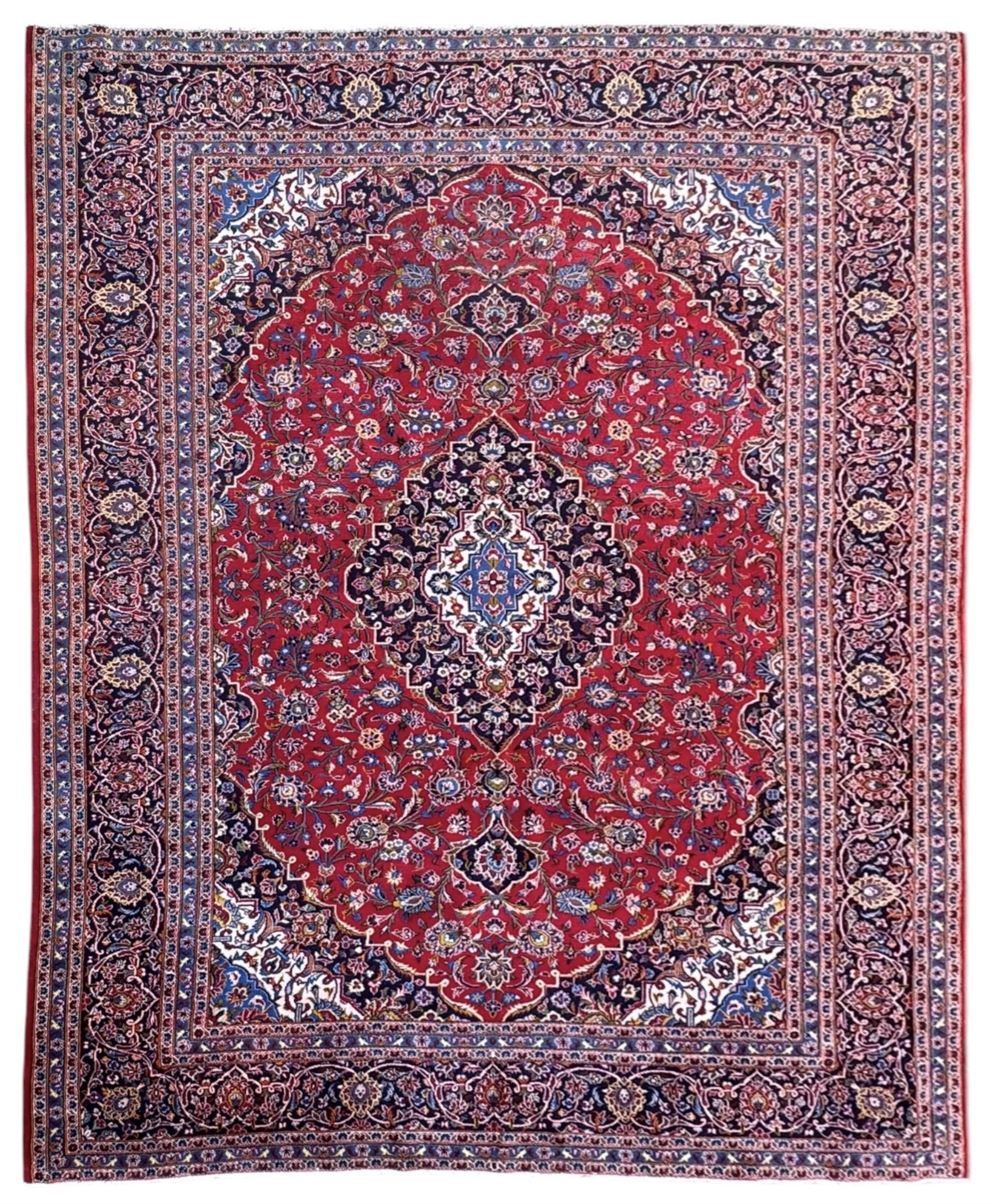 Large Kashan crimson ground carpet, central indigo medallion with stylised floral motifs, flanked by matching spandrels, bordered by multiple bands featuring repeating foliate and floral designs throughout