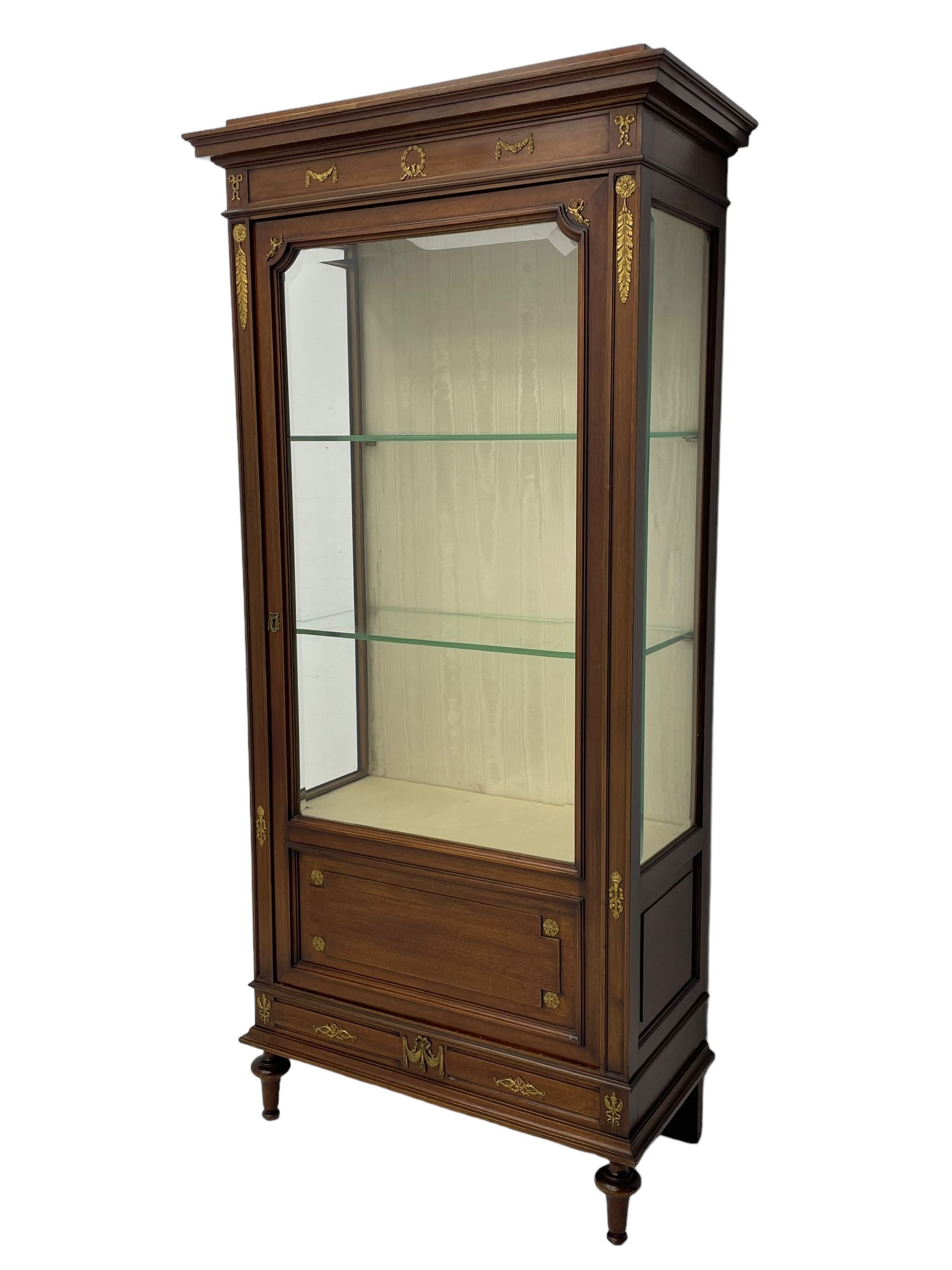 Early 20th century Louis XVI design mahogany vitrine display cabinet, projecting stepped cornice over recessed frieze decorated with gilt metal mounts of festoons and a bellflower wreath, fabric lined interior with two glass shelves, enclosed by bevel glazed door with lower panel, on two turned front feet 