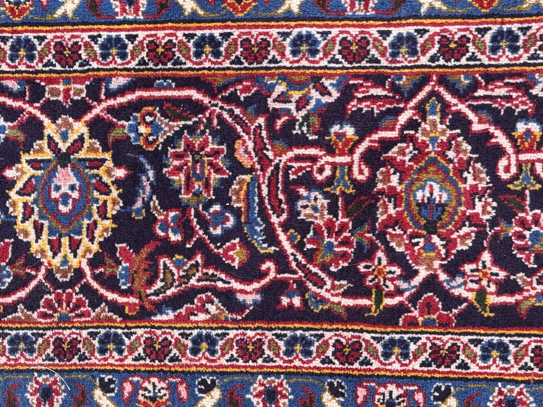 Large Kashan crimson ground carpet, central indigo medallion with stylised floral motifs, flanked by matching spandrels, bordered by multiple bands featuring repeating foliate and floral designs throughout