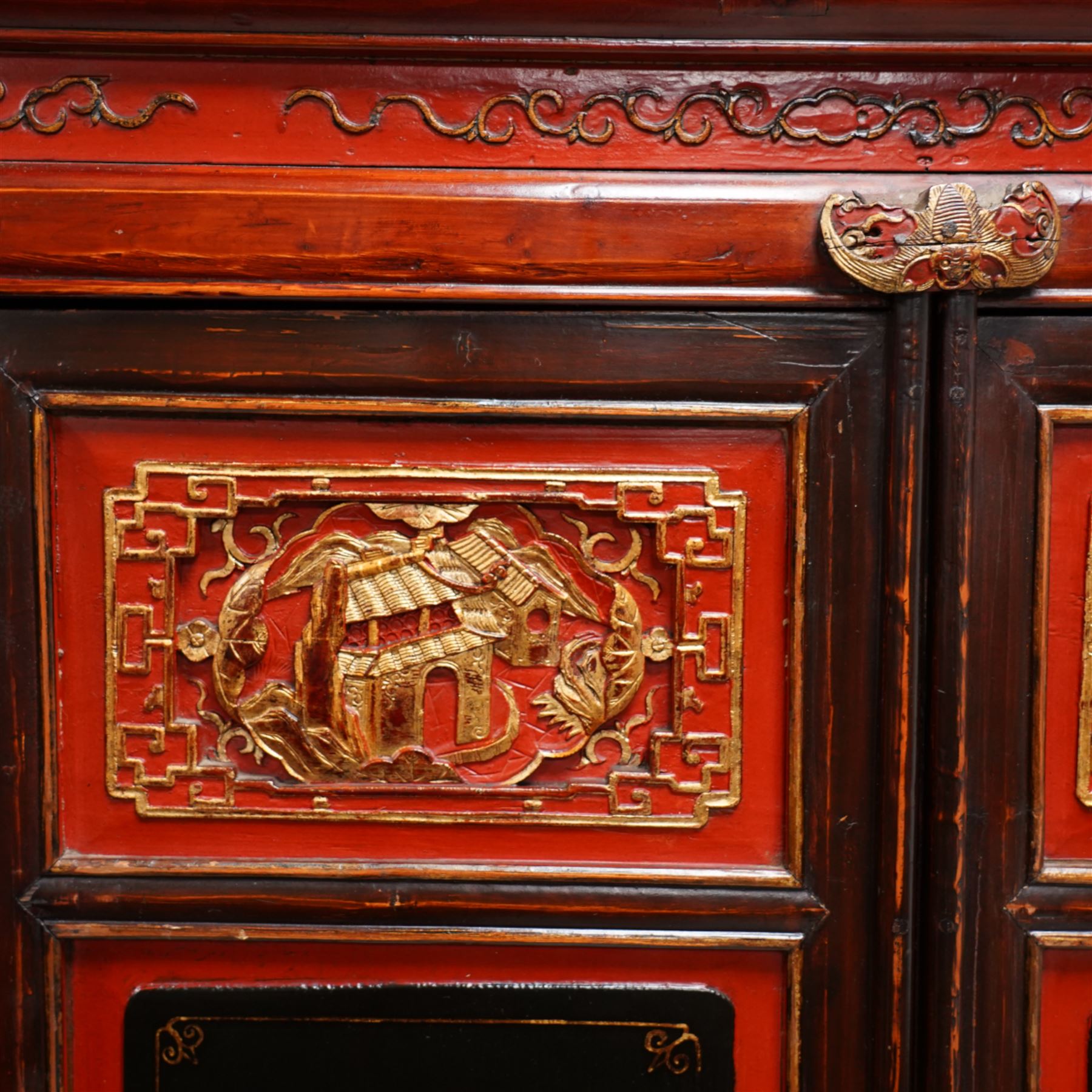 19th century Chinese red and black lacquered hardwood cabinet fitted with four panelled doors, the frieze decorated with scrolling gilt patterns over gold bat mounts surmounting the hinges, the black panels depicting traditional figural and floral scenes, the carved parcel-gilt panels with pagoda, pastoral and war scenes, the doors fitted with metal fish and pagoda handles, enclosing a single shelf and four internal drawers, the base compartment decorated with carved precious objects, raised on moulded square supports with pierced scrolling brackets