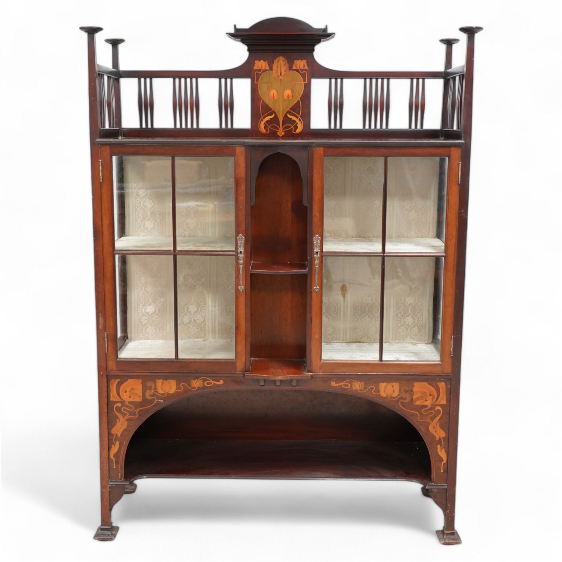 Shapland & Petter; Barnstaple (1854-1999) - Art Nouveau circa. 1900s mahogany display cabinet, raised shelf over panel inlaid with heart and stylised tulips with curled leaves, flanked by balustrade, enclosed by two astragal glazed doors, the lined interior fitted with two shelves, recessed central upright fitted with single shelf, arched lower section fitted with undertier, the spandrels inlaid with further stylised flower heads and trailing branches, on style supports terminating to moulded pad feet, the locks stamped 'S&B, B', retailed by 'Denby & Spinks, Leeds' plaque verso