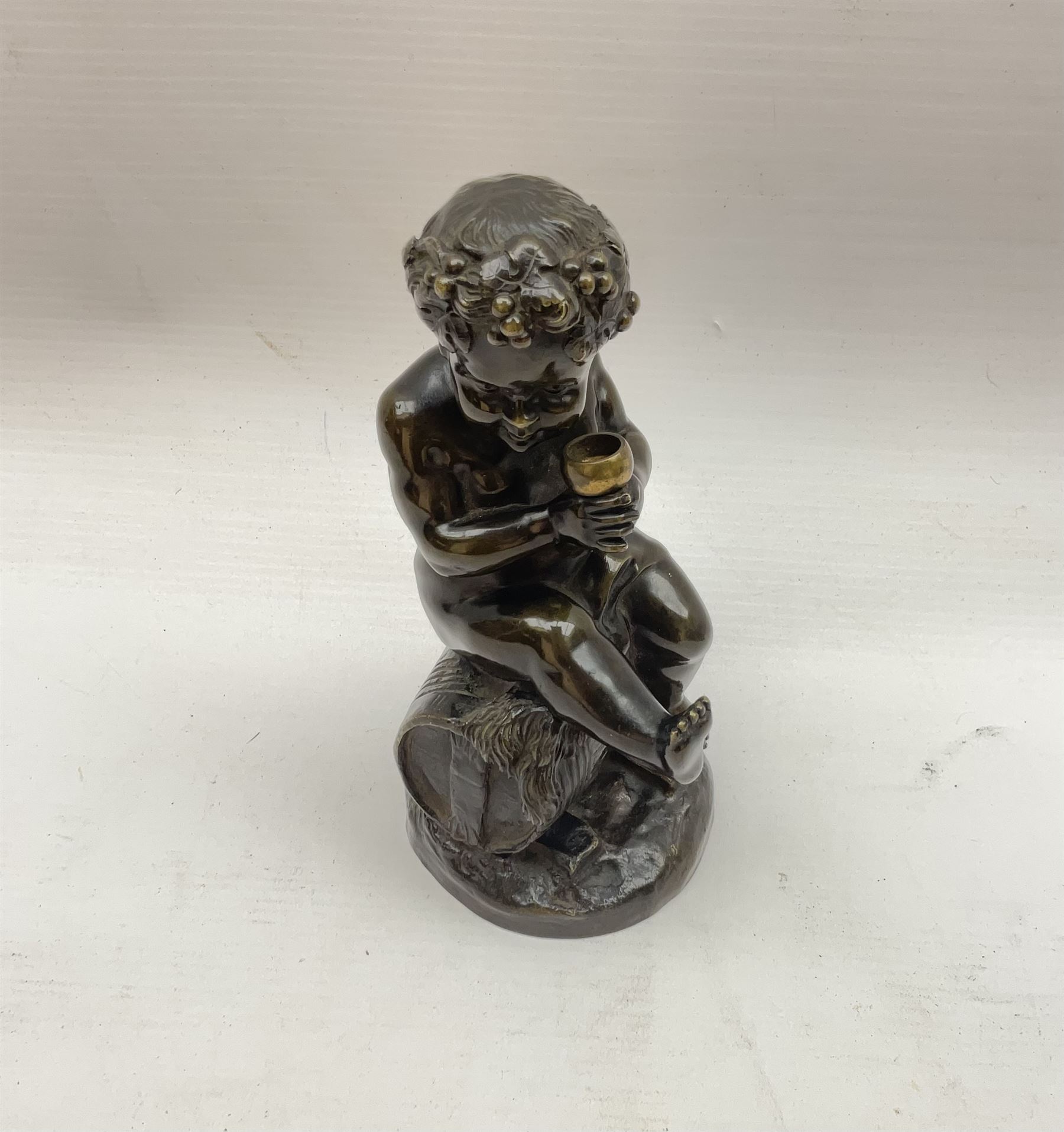 Bronzed metal model of a putti seated upon a barrel with goblet in hands, and fruiting vines adorning their head, H17.5cm