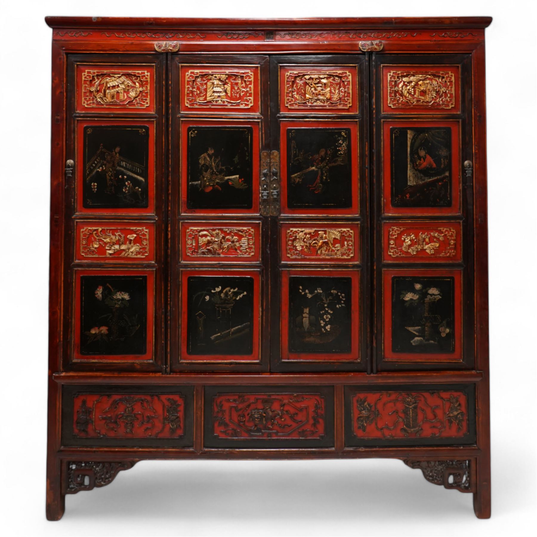 19th century Chinese red and black lacquered hardwood cabinet fitted with four panelled doors, the frieze decorated with scrolling gilt patterns over gold bat mounts surmounting the hinges, the black panels depicting traditional figural and floral scenes, the carved parcel-gilt panels with pagoda, pastoral and war scenes, the doors fitted with metal fish and pagoda handles, enclosing a single shelf and four internal drawers, the base compartment decorated with carved precious objects, raised on moulded square supports with pierced scrolling brackets