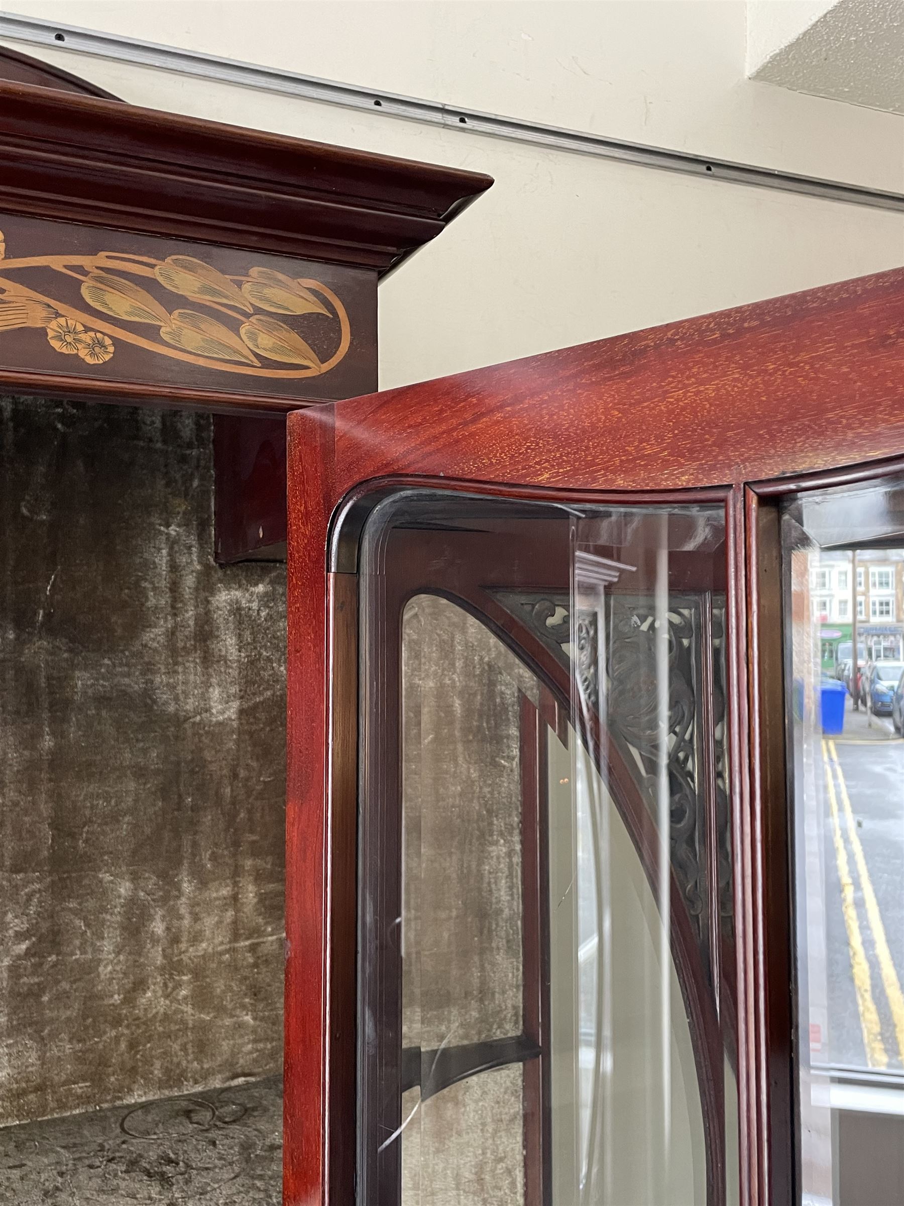 Art Nouveau period inlaid mahogany display cabinet, raised shaped and moulded pediment over projecting moulded cornices, the upper frieze inlaid with trailing branches and tulips, single astragal and bevel glazed door enclosed velvet lined interior fitted with shelves, flanked by curved glass panes decorated with pierced metal foliate spandrels, lower flanking shelves with mirror backs enclosed within shaped and pierced rails inlaid with flower heads, on tapering octagonal supports terminating to compressed pad feet, the lock stamped 'Salmon Bros, London' 