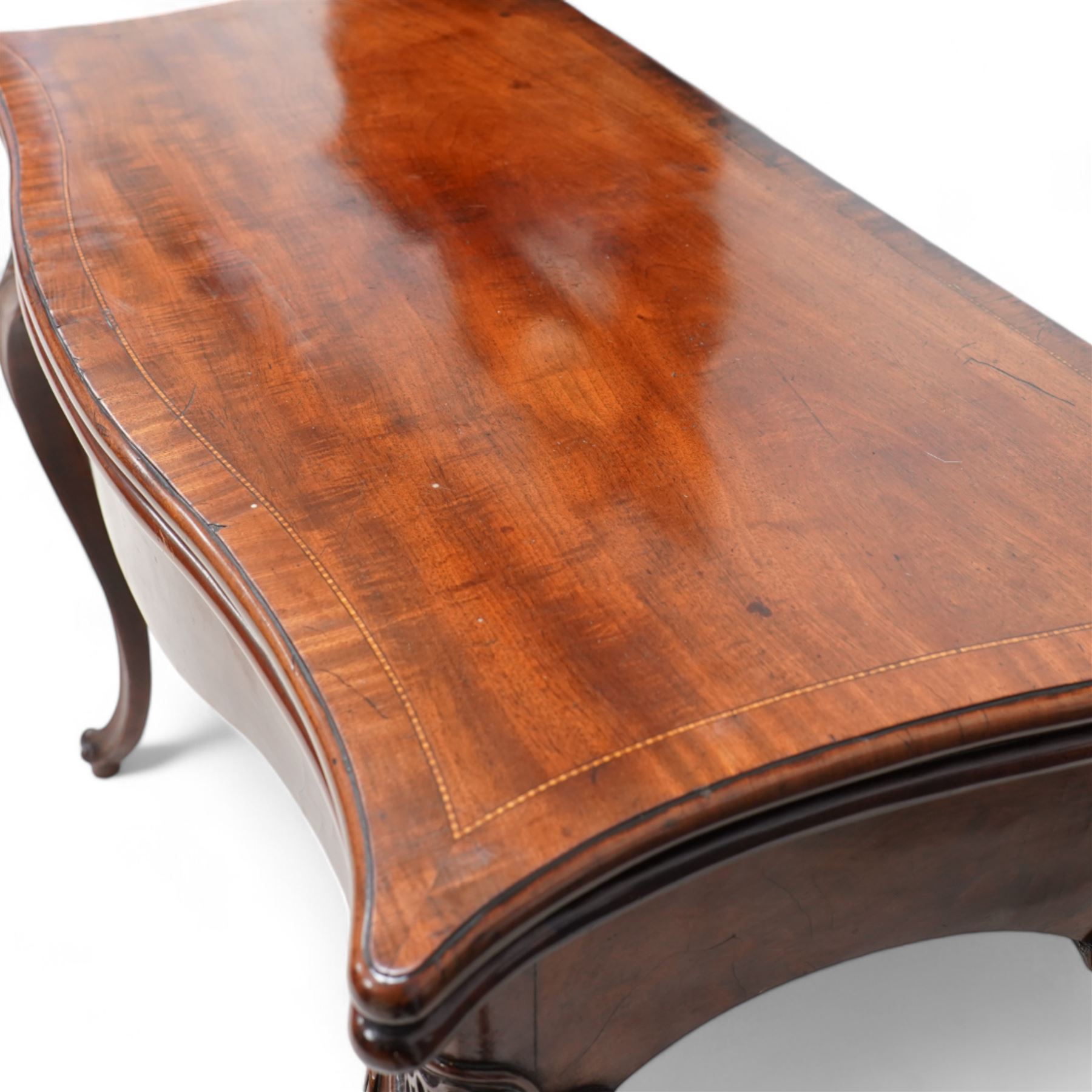 George III inlaid mahogany card table of serpentine form, the shaped rectangular fold-over top decorated with cross-banding and chequered inlay, opening to reveal a baize lined interior, the shaped apron over moulded cabriole supports with scroll and scallop carved knees, on scroll feet