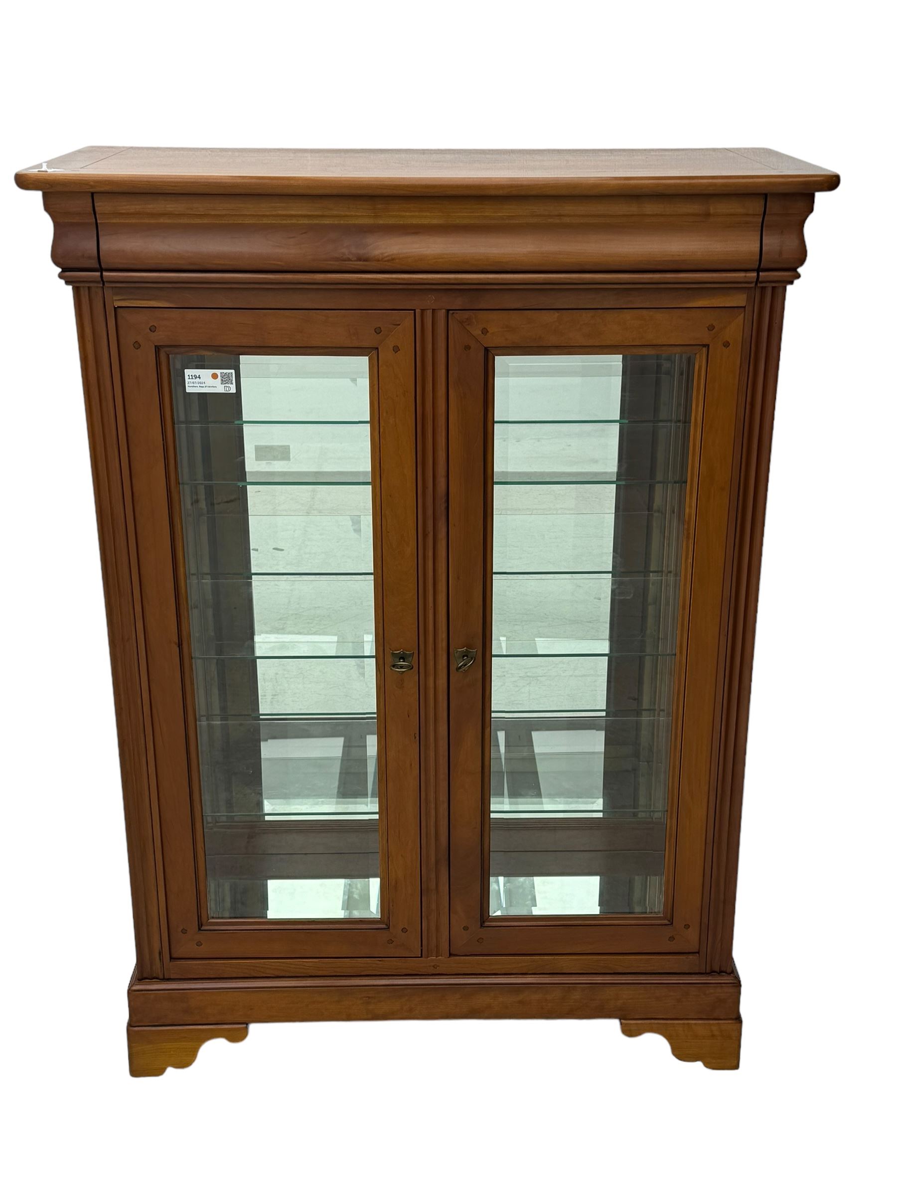 20th century cherry wood display cabinet, projecting cornice over two glazed doors with bevelled panes flanked by fluted support columns, enclosing three adjustable glass shelves with mirror-back surface, supported by shaped bracket feet