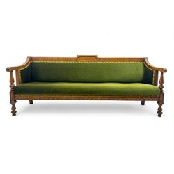 Late Victorian walnut framed sofa, rectangular backrest with carved detailing and central motif, upholstered in olive green fabric with brass studded trim and patterned front rail, scrolled fluted arms supported by turned uprights terminating in bun feet