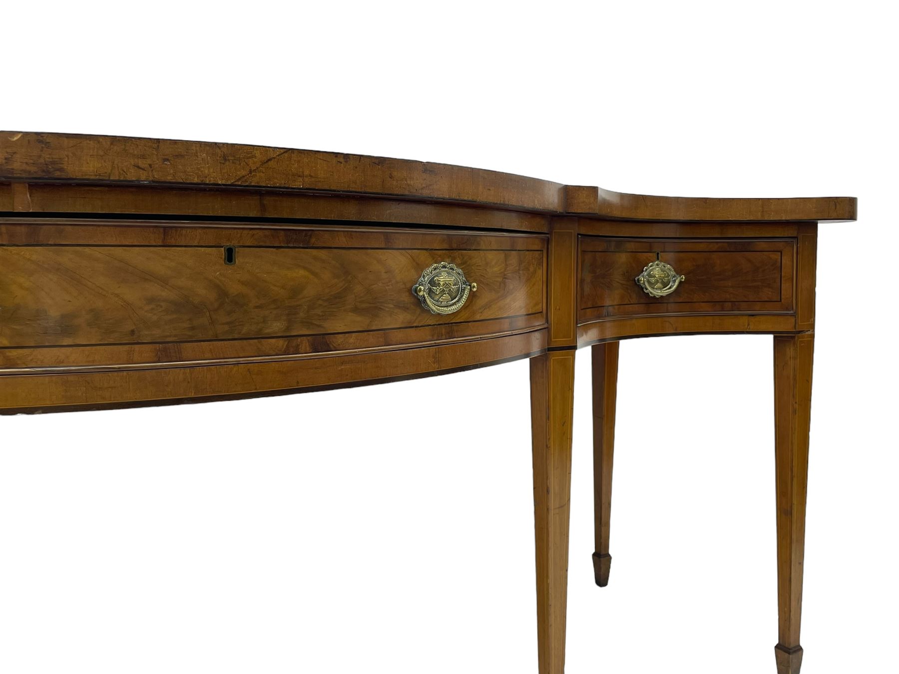 Large George III mahogany serpentine serving table, shaped top with crossing banding and stringing, the frieze fitted with three cock-beaded drawers, oval pressed brass handle plates decorated with urns, on square tapering supports with spade feet 